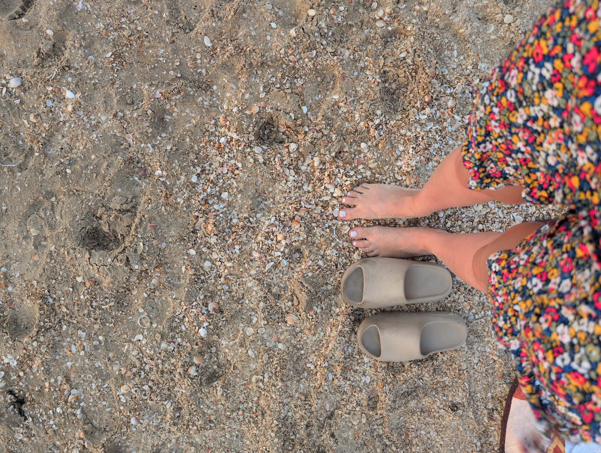 In Palmachim, Israel, a pair of beige slides lies on a sandy beach littered with small seashells. A person's bare feet, with white-painted toenails, stand in the sand next to the slides, while the lower portion of their legs and a colorful floral skirt are visible in the foreground.