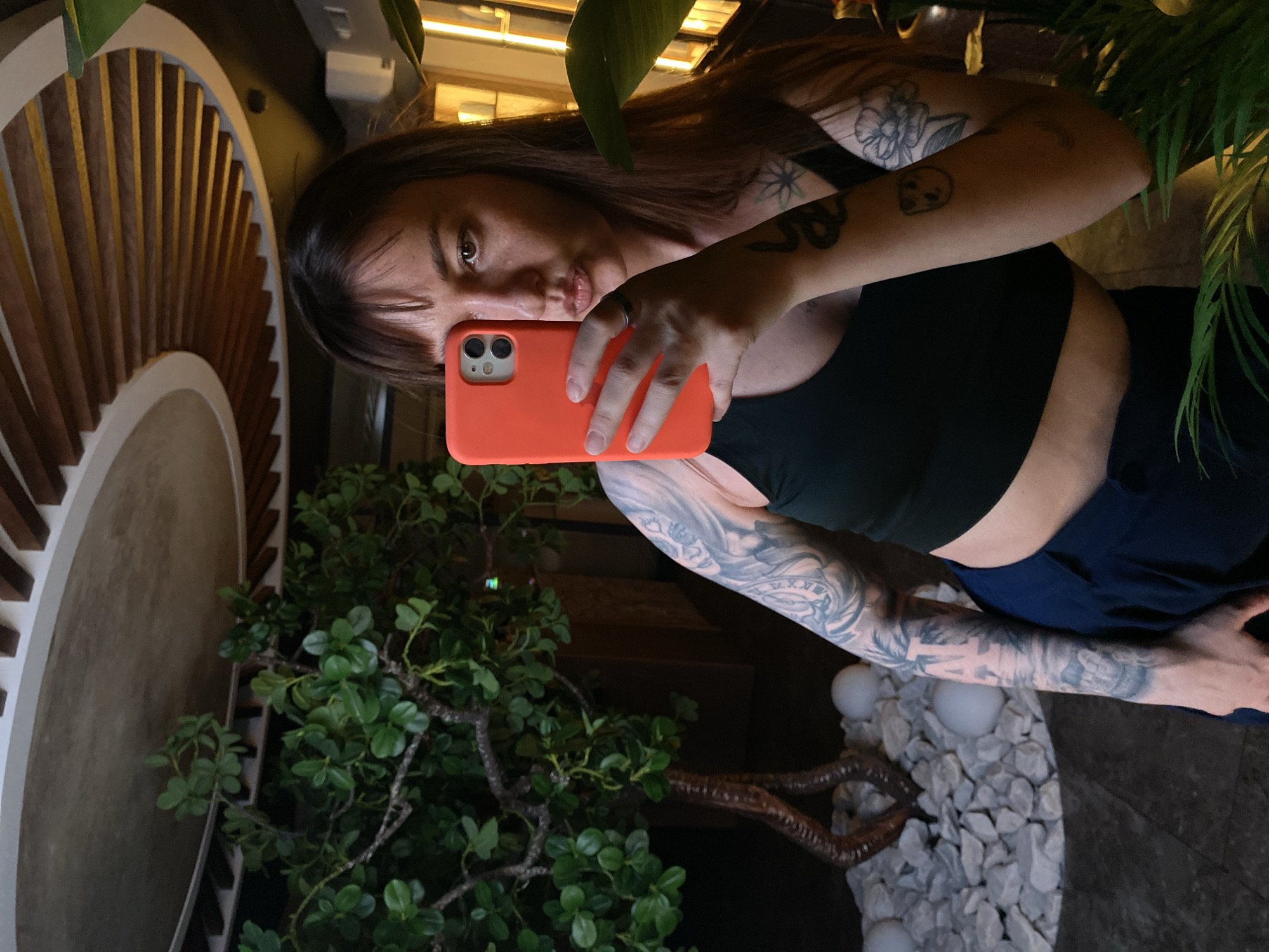 A woman with tattoos on her arms is taking a selfie using a bright orange phone. She is wearing a dark-colored top and dark bottoms. The background includes foliage, a circular structure with vertical slats, and a decorative arrangement of white stones and spheres. While a precise location is difficult to determine without additional context, the image does not contain information to confirm that it was taken in the city of Votkinsk, Russia.
