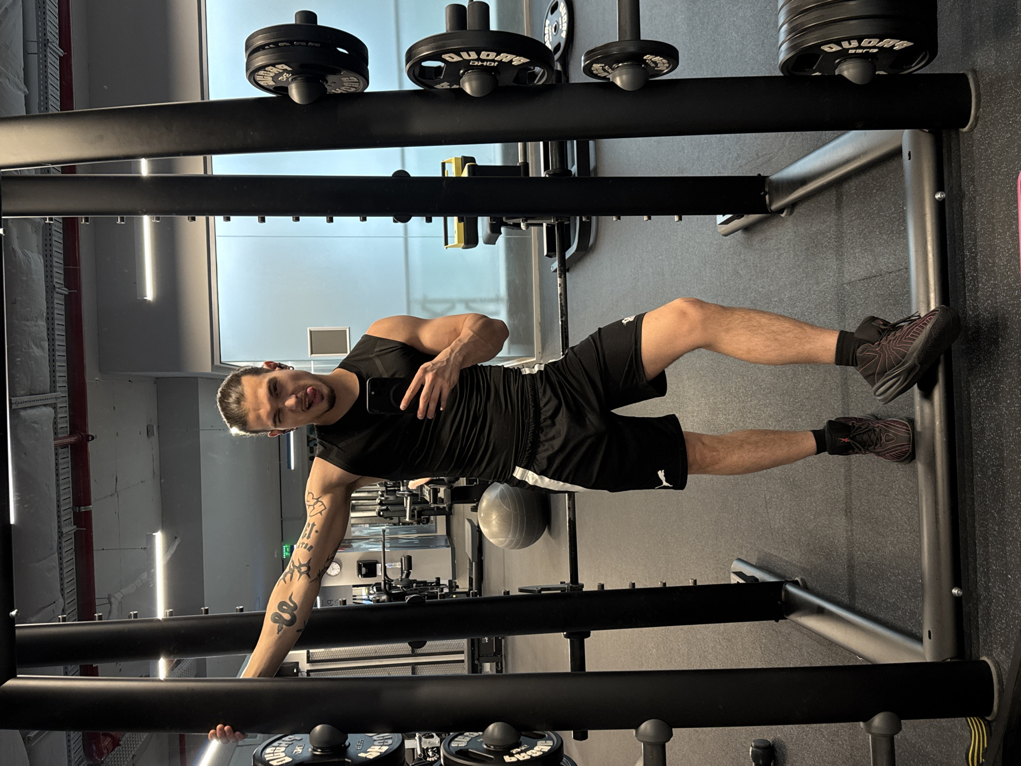 A young man with dark hair tied back in a bun, wearing a black sleeveless shirt and black shorts with white stripes, is taking a selfie in a gym. He has tattoos on his left arm and is sticking his tongue out, winking. He is standing in front of a black power rack with weight plates on it. In the background, there are other gym equipment, including a large exercise ball and weight benches. The gym has a modern industrial aesthetic with exposed ceilings and fluorescent lighting. The floor is dark rubber matting. The scene appears to be set in a gym in Sofia, Bulgaria, suggested by the presence of specific gym equipment brands and architectural styles common in the city.