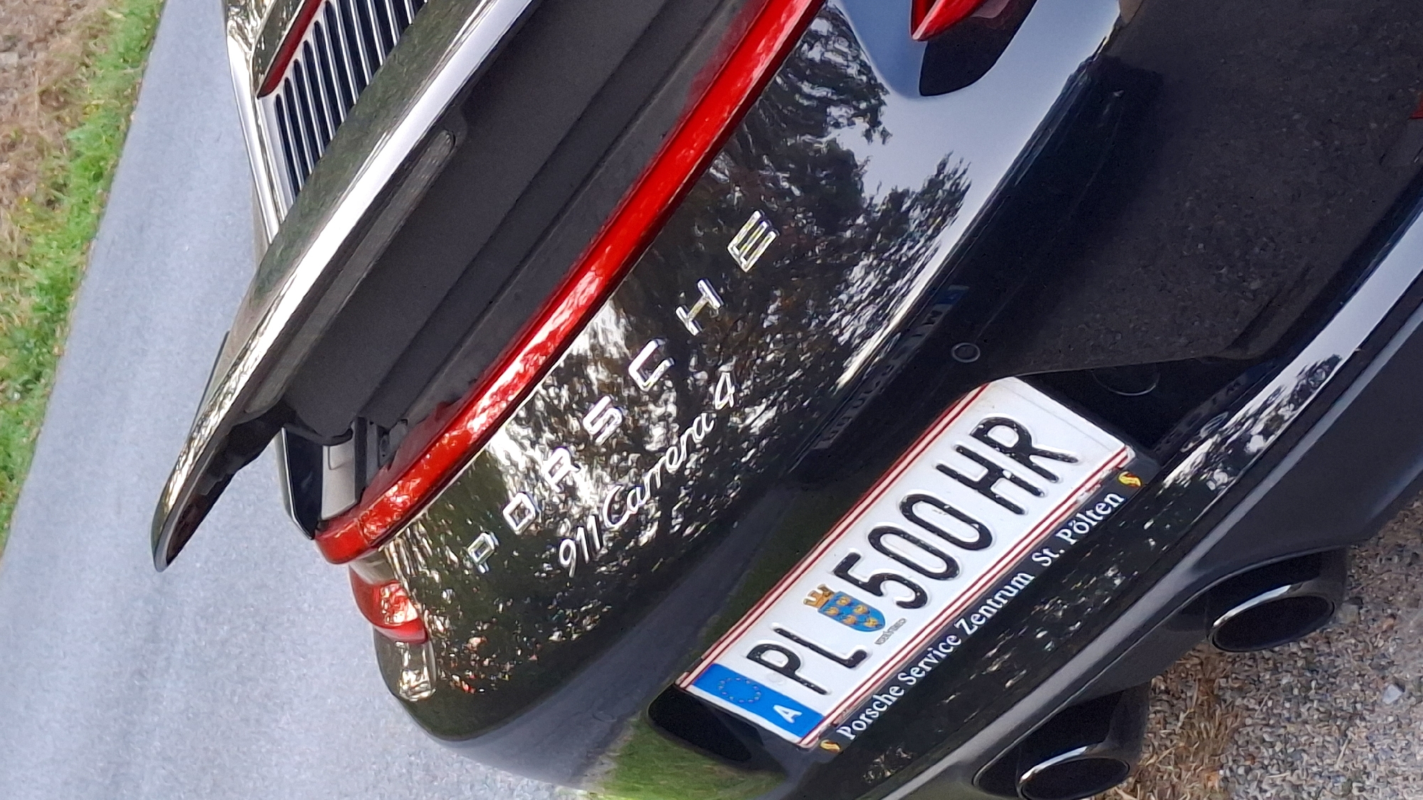 The image shows the rear of a black Porsche 911 Carrera 4. The car is parked outdoors on a paved surface, likely a road or parking area, with a grassy verge visible in the background. The license plate is visible and reads "PL 500 HR" with "A" in a blue European Union section. Below the license plate, text indicates "Porsche Service Zentrum St. Pölten". The car has visible red accents on its rear spoiler and taillights. Two black exhaust pipes are visible at the bottom of the image. The scene appears to be in the municipality of Rosenburg-Mold, Austria, as suggested by the presence of a Porsche service center in St. Pölten, a city within the region. There are no people or other significant objects visible in the immediate vicinity. The lighting suggests it is daytime, possibly overcast due to the reflections on the car's surface.
