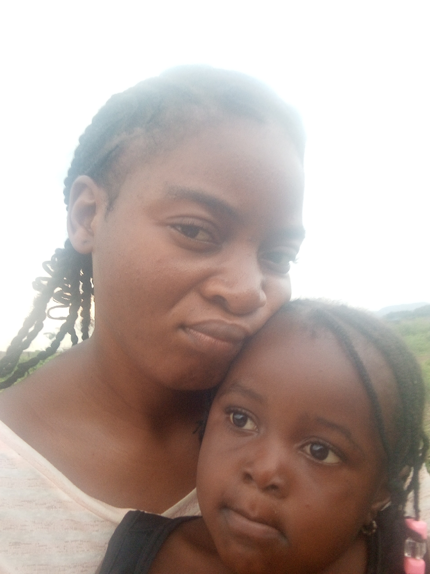 In the city of Serti, Nigeria, a selfie is captured, featuring a woman and a young child. The woman, positioned on the left, has her hair styled in braids that hang down her left side. She is looking towards the camera with a slight smile. The child is nestled against the woman on the right side of the frame, with their head resting on the woman's shoulder. The child's face is turned towards the camera, with their eyes looking slightly off to the side. The background of the image appears to be an outdoor setting, with a hint of green foliage visible in the distance. The lighting suggests it is daytime, possibly with some overcast conditions.