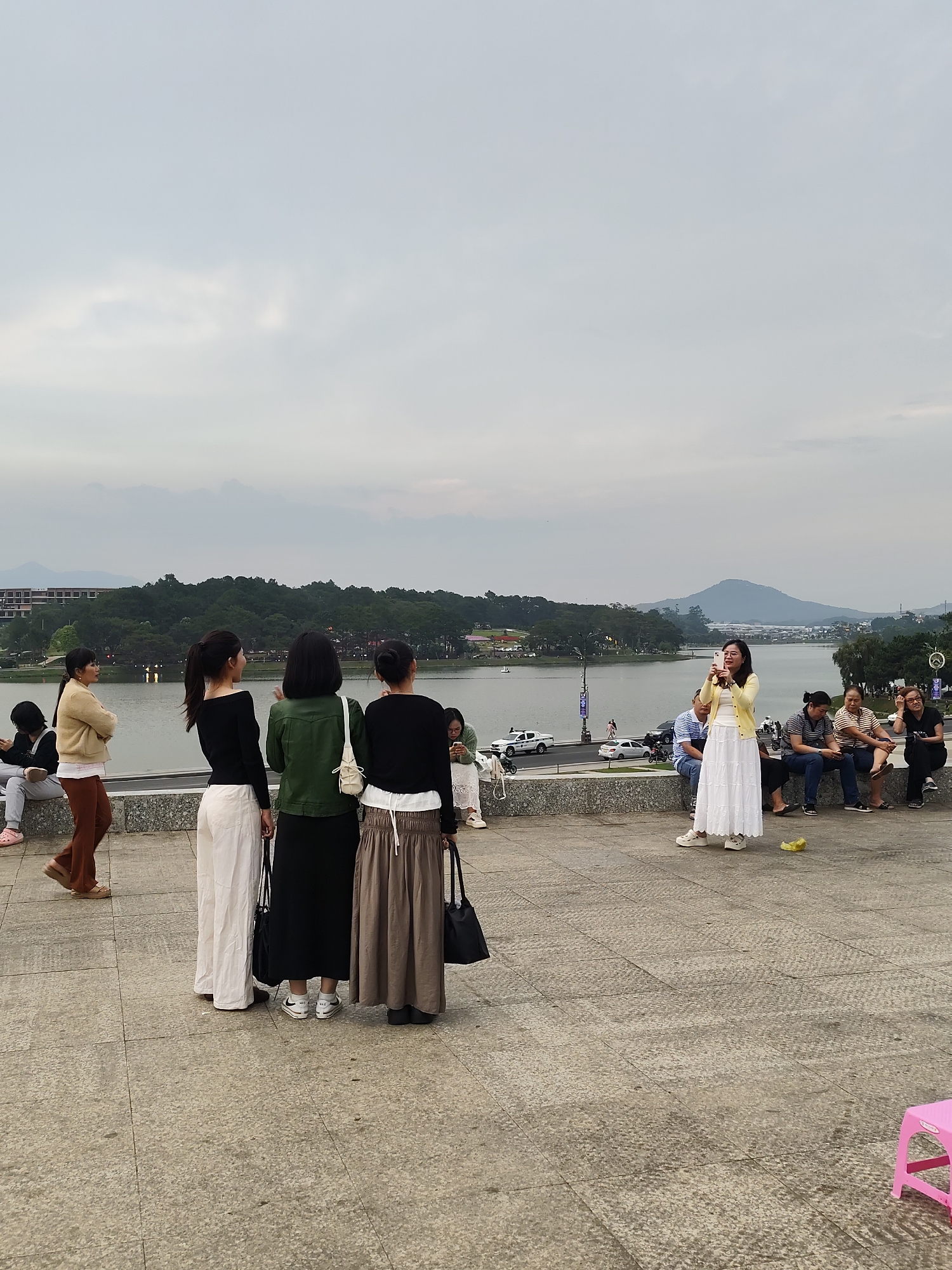 The image captures a scenic view over a lake with forested hills and distant buildings, characteristic of Da Lat, Vietnam, under an overcast sky. In the foreground, on a paved outdoor area, several people are visible. A central group of four women stands, with three of them facing away from the camera, observing the expansive view. Their attire is casual, featuring long skirts or wide-legged pants in neutral tones like white, black, and brown, paired with dark tops and a dark green jacket on one. Two of these women wear white sneakers, with the "Nike" logo visible on one pair.
To the right of this group, a woman in a white maxi skirt and a bright yellow cardigan stands, holding a smartphone up to take a photo of the lake and hills. Further to the left, another woman in a yellow cardigan and brown flared pants walks across the frame, looking towards the left. Along a low concrete wall that separates the paved area from a road below, several individuals are seated, relaxing and enjoying the scenery. Cars are visible on the road. A small, bright pink plastic stool is partially visible in the bottom right corner, and a crumpled yellow wrapper lies on the pavement near the woman in the white skirt. The grey, cloudy sky suggests either an overcast day or late afternoon, casting a soft, diffused light over the tranquil scene.