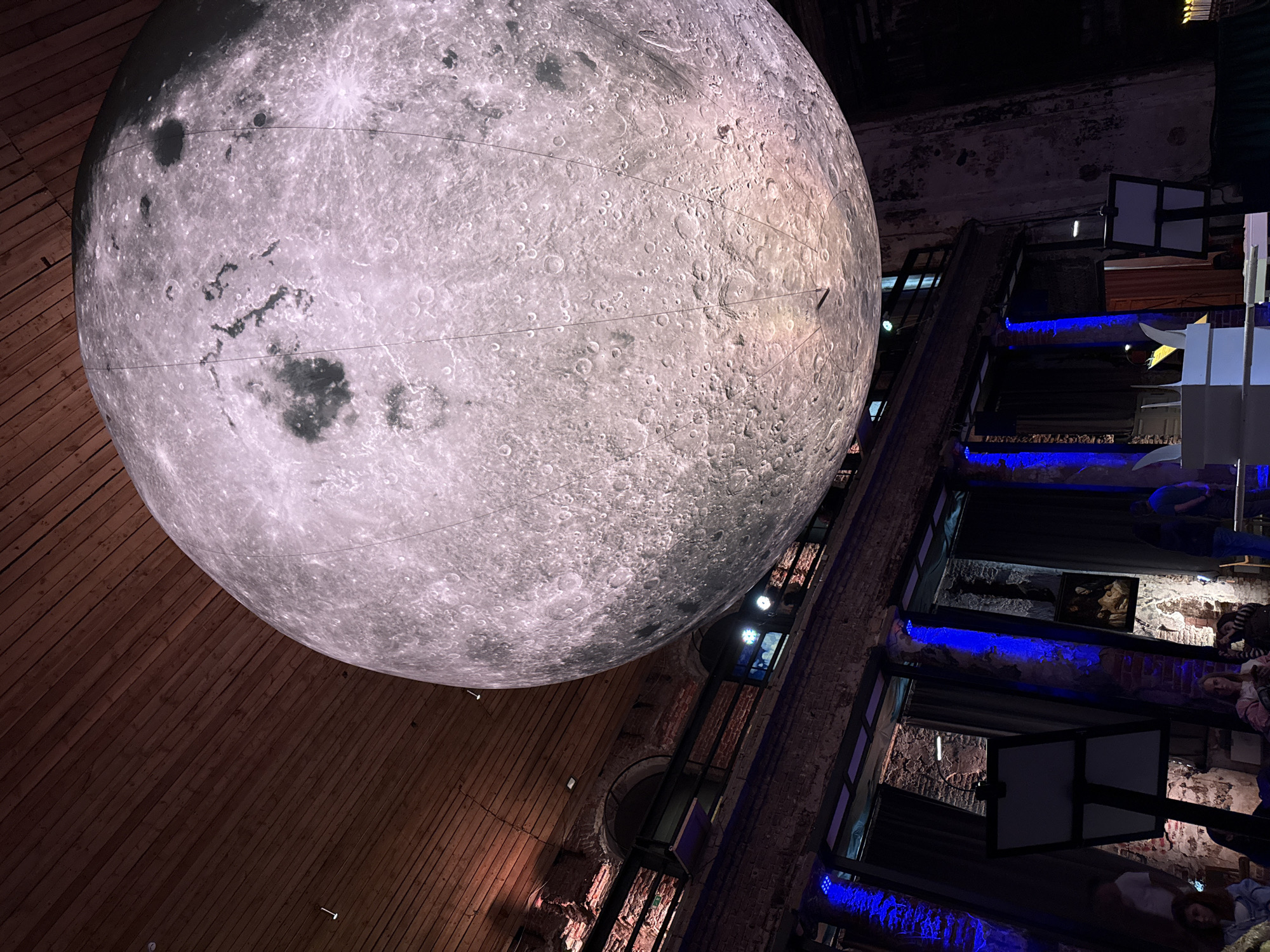 The image depicts a large, illuminated replica of the moon suspended from the ceiling of a dimly lit interior space. The replica is detailed, showing craters and surface features. Below the moon, the environment appears to be a venue with exposed brick walls, wooden beams, and draped curtains. Blue lighting illuminates some of the architectural features. Several people are visible in the lower portion of the image, seated and standing, some appearing to be attending an event. Two easels with blank white boards are positioned in the foreground. This scene is located within the Museum of the World Ocean in Kaliningrad, Russia.