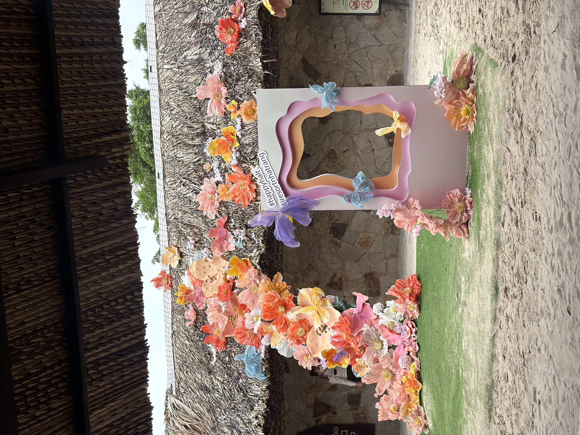 The image shows a decorative floral display and a photo booth setup in front of a thatched-roof structure with a stone wall behind it. The photo booth is a pastel pink and orange frame adorned with fabric flowers and bows. A string of hashtags, "#happyhalt #resortnhatrang", is visible on the photo booth. A woman wearing a face mask and a light-colored shirt is partially visible in the background to the left, standing near an entranceway. The foreground consists of sand and a patch of green artificial grass. The overall scene suggests an outdoor recreational area or resort in Nha Trang, Vietnam.