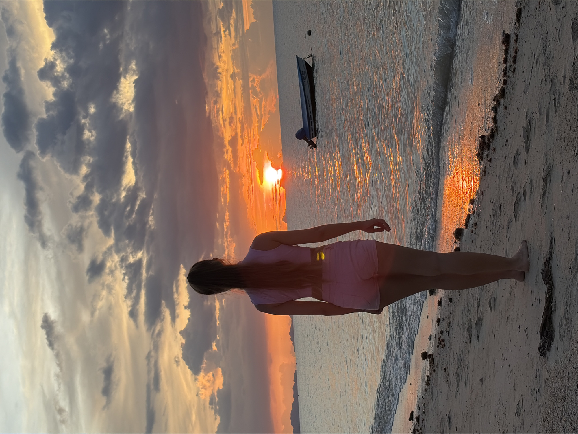A young woman stands on a sandy beach, facing the ocean and a vibrant sunset over the water. She has long brown hair pulled back in a ponytail and is wearing a pink crop top and pink shorts. The sky is filled with dramatic clouds colored in shades of orange, yellow, and gray. In the distance, islands are visible on the horizon. A small boat with an outboard motor is moored in the calm water to the right of the frame. The waves gently lap at the shore, reflecting the warm hues of the sunset. The scene appears to be located on a beach in Baan Thong Po, Thailand.