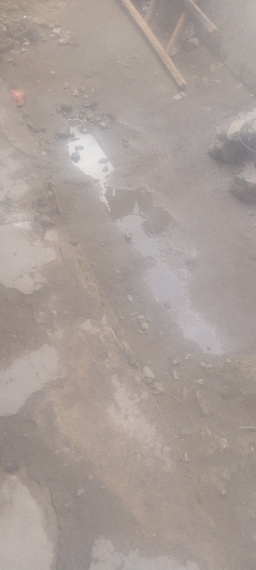 The image shows a muddy, debris-strewn ground with several puddles of standing water. The location is potentially Sabon Tasha, Nigeria, although this cannot be confirmed based on the image alone. Scattered rocks and construction-related materials are visible. A small orange container rests near one edge of the frame. Pieces of wood, possibly used for scaffolding or construction, are in the upper right corner. The overall impression is that of an unfinished or disrupted construction site after rainfall or a period of wet weather. There are no people visible in the image.