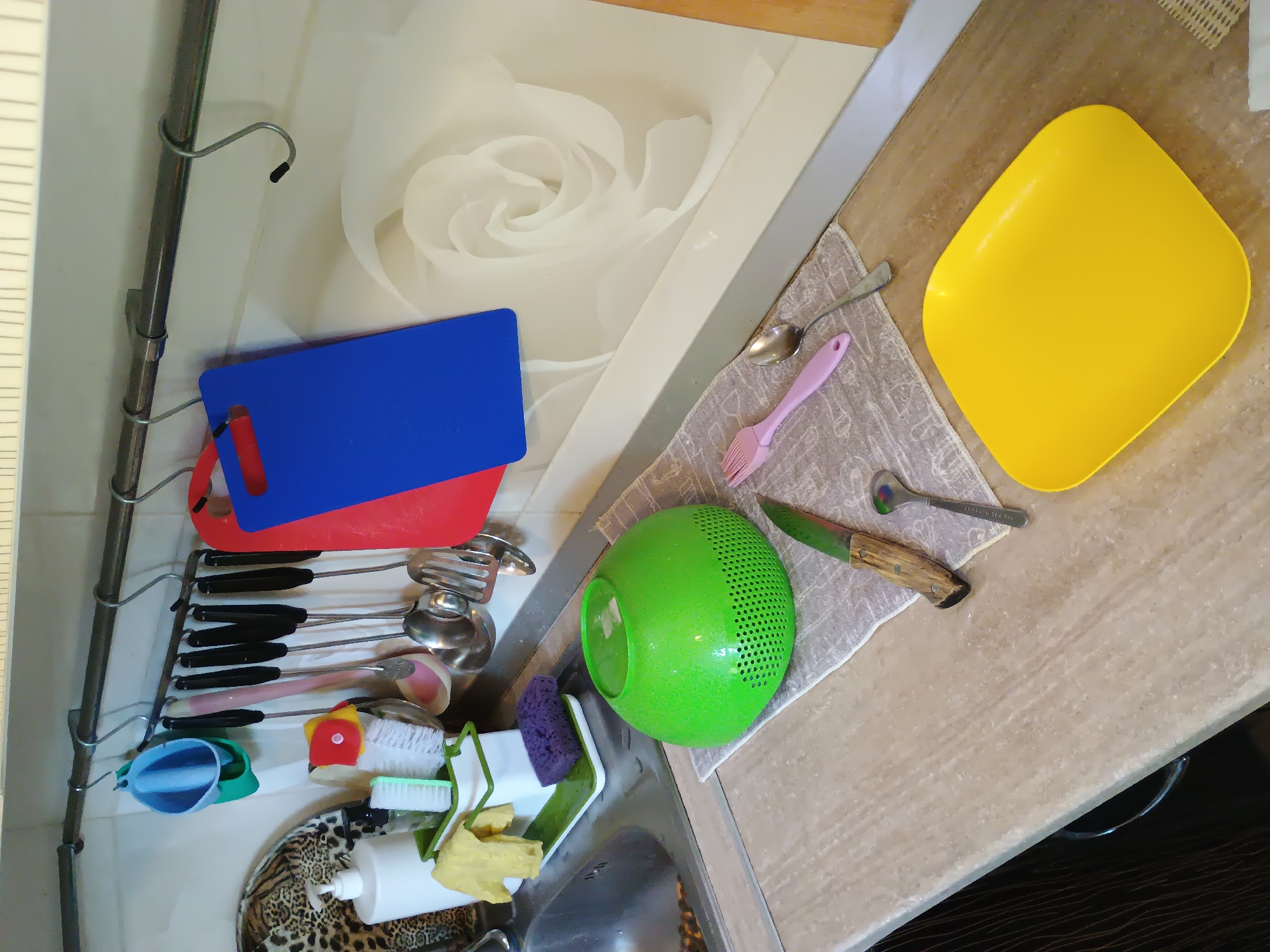 This is a top-down, slightly angled, well-lit, indoor shot of a kitchen sink area. The scene features various kitchen utensils and cleaning supplies. Prominently displayed are a bright yellow square plate, a green colander, a knife with a wooden handle, a pink fork, and several metal spoons. To the left, two cutting boards, one red and one blue, are hung on a metal rack along with a collection of ladles and other serving utensils. Below them, a blue plastic cup and a green scoop are visible, along with cleaning brushes and a yellow sponge. A white pump bottle sits on a leopard-print surface near the sink. In the background, a large, stylized white rose is depicted on a wall or backsplash. The setting appears to be a home kitchen, likely in Zaporizhzhia, Ukraine, given the location context. The lighting suggests it could be daytime, and there are no indications of specific weather or emotional cues. There is no visible text in the image.