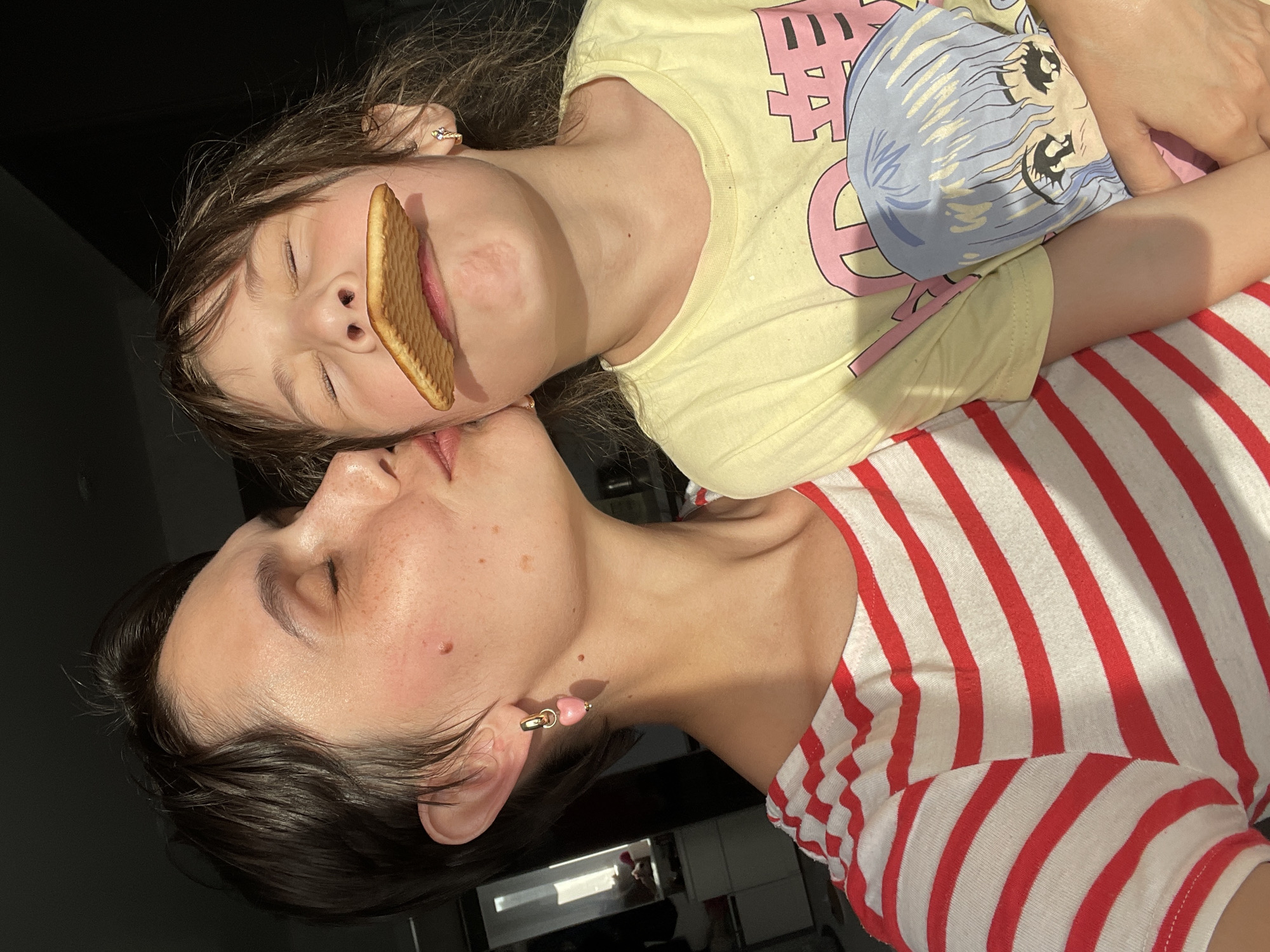 A woman and a child are lying down side by side, facing upwards towards the camera. The child, who is wearing a yellow shirt with an anime character, has a large, rectangular cookie positioned over their mouth. The woman is wearing a striped red and white shirt. The background appears to be an indoor setting with a window or reflective surface visible. The location and city cannot be determined from the visual information provided.