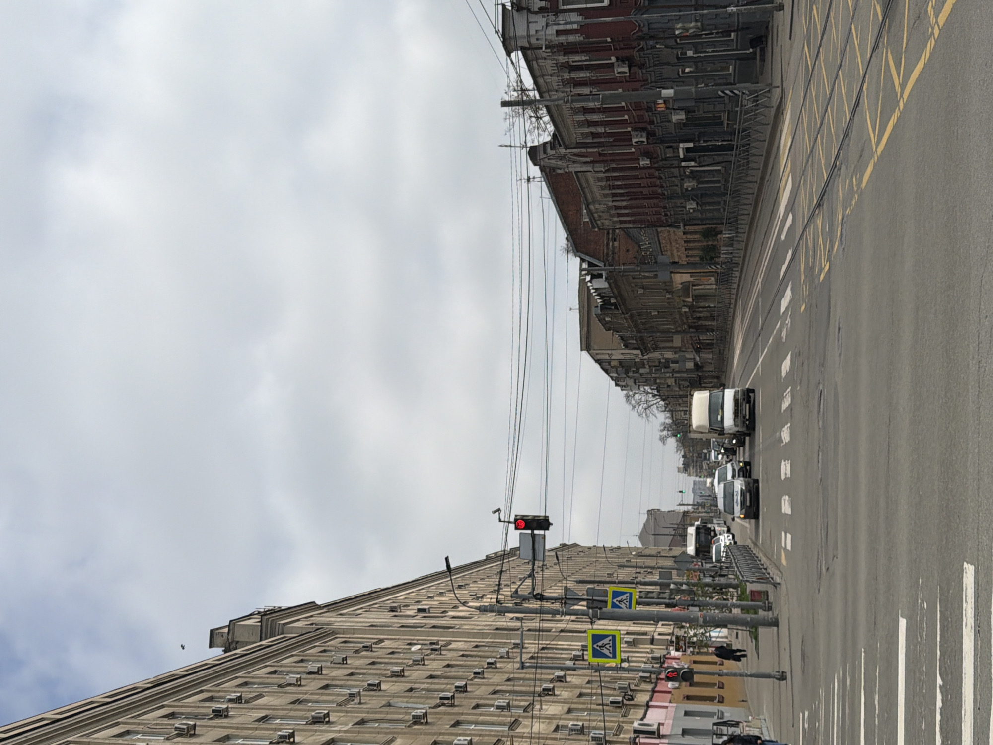 The image depicts a street in Rostov-on-Don, Russia. The view is along a straight avenue lined with buildings. On the right, a multi-story brick building with intricate architectural details stands next to a road marked with yellow stripes. On the left, a tall building with multiple rows of windows and numerous air conditioning units is visible. Overhead, power lines crisscross between the buildings. The road is paved and features pedestrian crossings marked with white stripes. Several vehicles, including vans and cars, are travelling down the street. Traffic lights are visible, with at least one showing a red light. There are also pedestrian crossing signs with the image of a pedestrian on them. The sky is overcast.