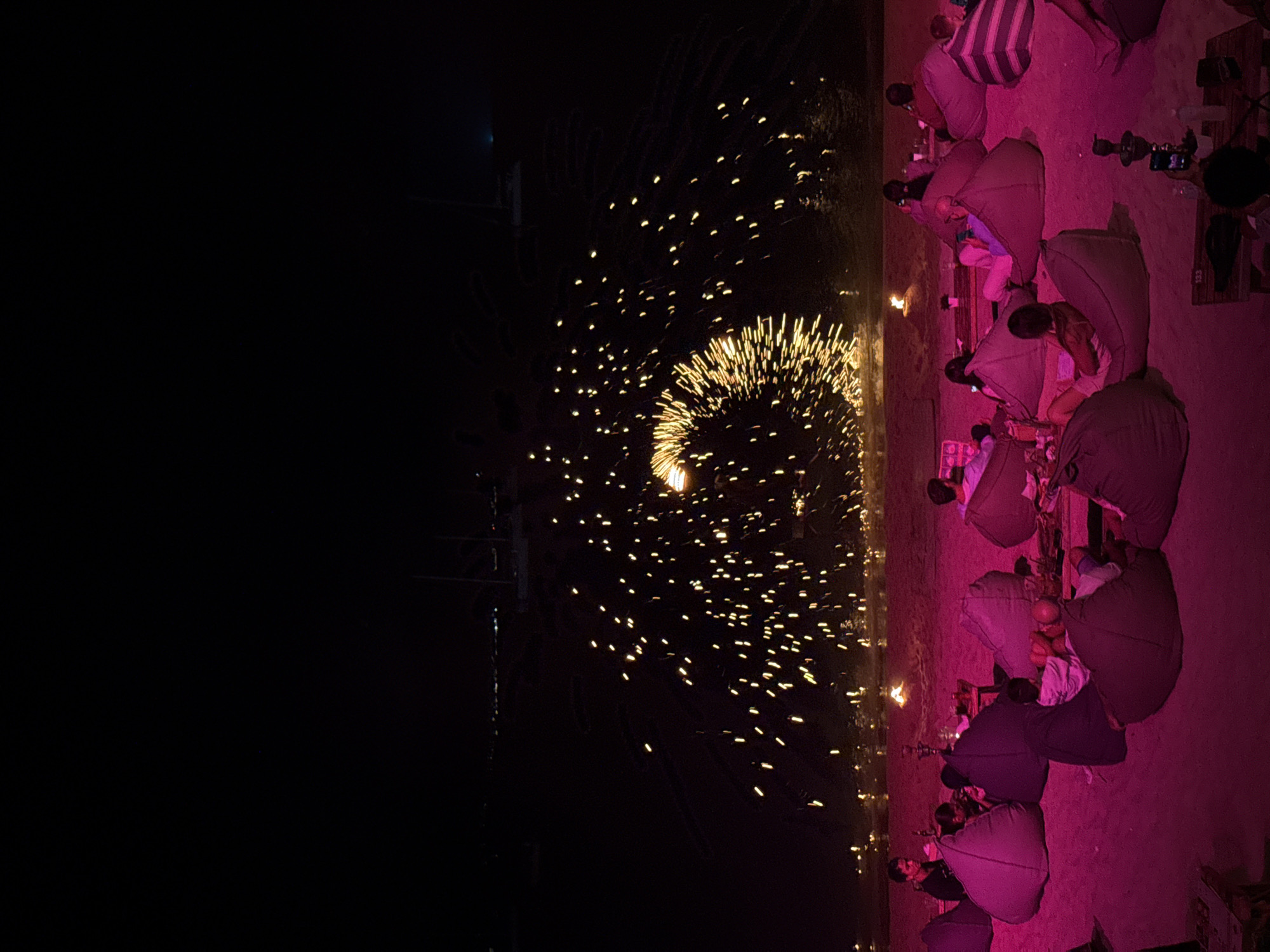 The image depicts a beach at night, likely in Ko Samui District, Thailand, with a fire performance taking place over the water. The scene is illuminated by pink ambient lighting and the sparks from the fire performance. Several people are seated on beanbag chairs on the sandy beach, facing the water and the performance. The sky is dark, with some faint lights visible in the distance, possibly from boats or a coastline. The fire performance is creating a circular pattern of bright, golden sparks. The overall atmosphere appears relaxed and festive.