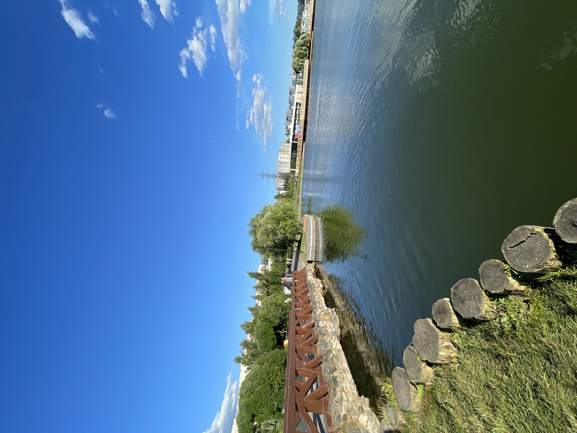 This is a bright, sunny day in a park setting, likely Moscow, Russia. The image shows a wide, calm body of water, possibly a river or lake, with ripples reflecting the blue sky and scattered white clouds. Along the water's edge, there is a landscaped area with a stone embankment topped by a wooden railing. Beyond the railing, there are trees, green grass, and several people can be seen walking and sitting, enjoying the park.
In the background, a city skyline is visible, with buildings and a tall electrical pylon suggesting an urban environment. The water appears to be a dark green color, and the sunlight creates a strong reflection on its surface. The foreground features a rustic edge made of cut logs, interspersed with grass and some small pink flowers. The overall atmosphere is peaceful and recreational, typical of a park on a pleasant day. The time of day appears to be late morning or early afternoon due to the clear blue sky and strong sunlight. No visible text is present in the image.
