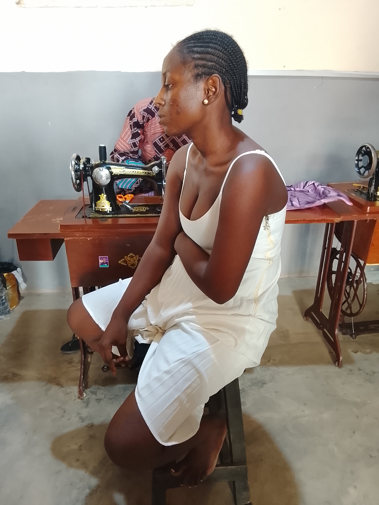 This image captures a young woman seated on a stool in what appears to be a tailoring workshop or training center in Akwanga, Nigeria. She has dark skin and braided hair, wearing a light-colored (possibly white) spaghetti-strap dress. She is looking to her right, away from the camera, with a neutral expression.
In the midground, a prominent vintage-style sewing machine with a dark wooden table is positioned to the woman's left. Fabric with a striped pattern and a pair of orange-handled scissors are on this table. A small, unreadable sticker is visible on the side of the table. Another similar sewing machine is partially visible on a wooden table to her right. In the background, behind the woman, a person wearing a patterned garment is indistinctly seen. Fabric, possibly purple, lies on the table to the woman's right.
The setting is indoors, featuring a concrete or cement floor and two-toned walls with a grey lower section and a lighter upper section. Bags or bundles are visible on the floor in the left background. The overall scene suggests an active, functional environment dedicated to sewing or garment production. The time appears to be daytime. No discernible text is visible on any objects.