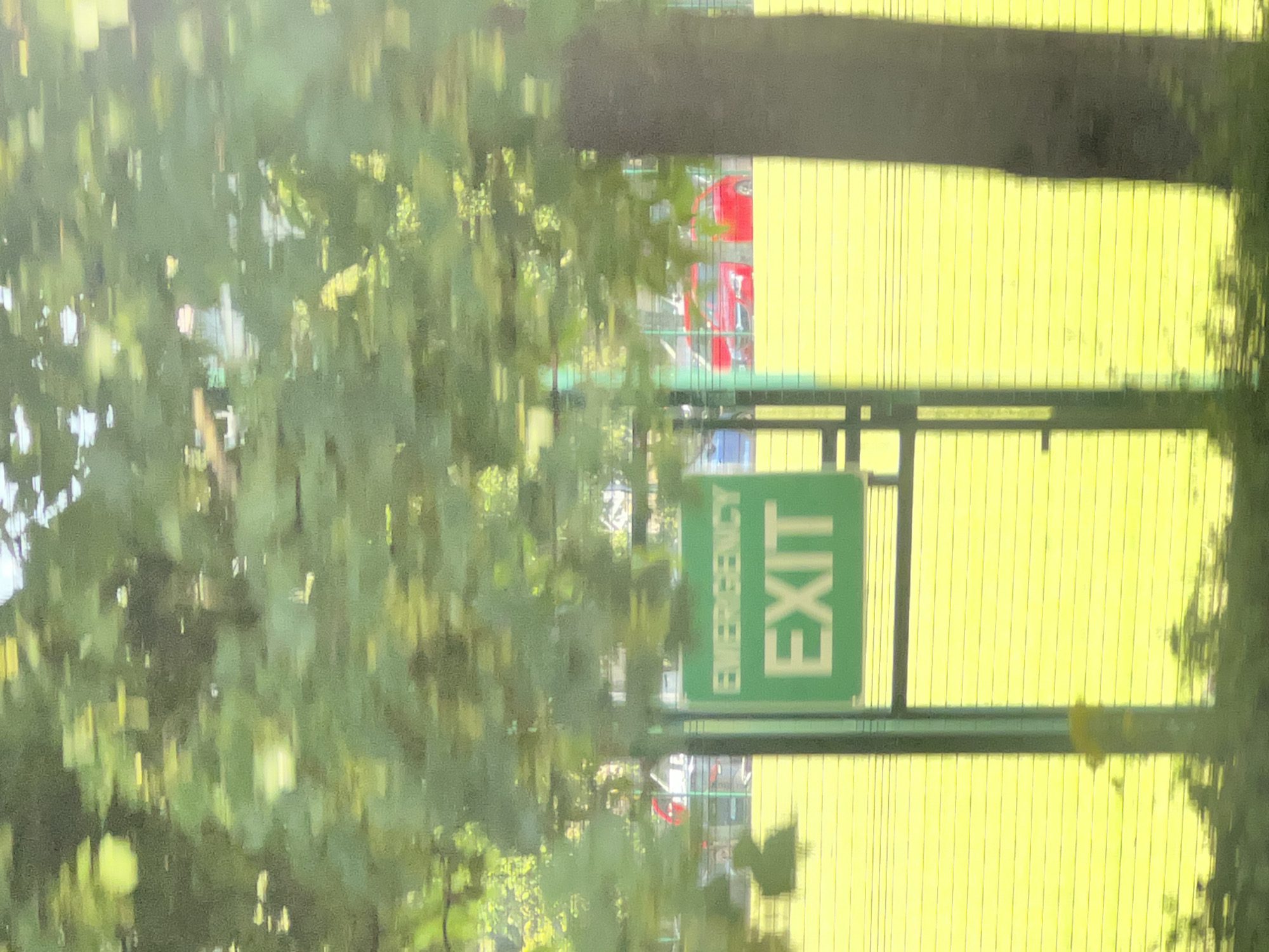 The image shows an "EMERGENCY EXIT" sign in green and white, positioned in front of a bright yellow field, likely a sports pitch. Behind the sign, two red cars are visible, parked on a road or car park. The scene is blurred due to motion, suggesting the image was taken from a moving vehicle. Trees with green foliage dominate the background. The location is specified as Letterkenny, Ireland.