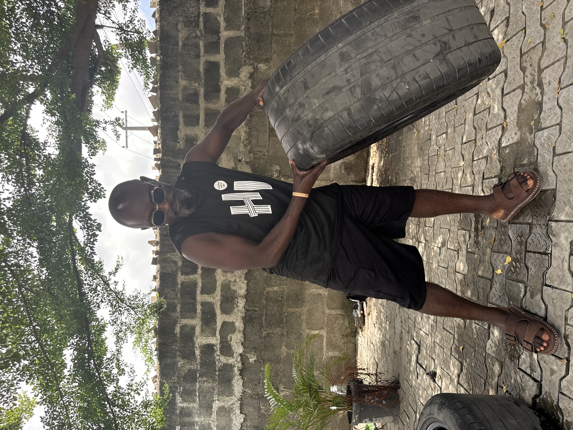 An outdoor, brightly lit, medium shot shows a dark-skinned man in what appears to be a compound or backyard. The man, with a beard and wearing sunglasses, a black sleeveless t-shirt, and black shorts, is actively engaged in lifting or holding a large, heavy black tire. He wears brown sandals on his feet.
The man's black sleeveless shirt features a white circular patch with visible text "My future is..." followed by a logo or design made of horizontal and vertical lines. A golden bracelet with Hebrew script is visible on his right wrist.
The setting includes a grey brick wall in the background, topped with an irregular edge, and a grey paved ground beneath his feet. Some sections of the paving appear wet, with scattered dry leaves. Two potted plants, one with green fern-like foliage and another with dried reddish branches, are positioned against the wall. Overhead, lush green tree foliage is visible against a bright, slightly overcast sky, with power lines and a utility pole faintly discernible in the distance. The scene suggests a warm, daytime environment in a residential or semi-urban area.