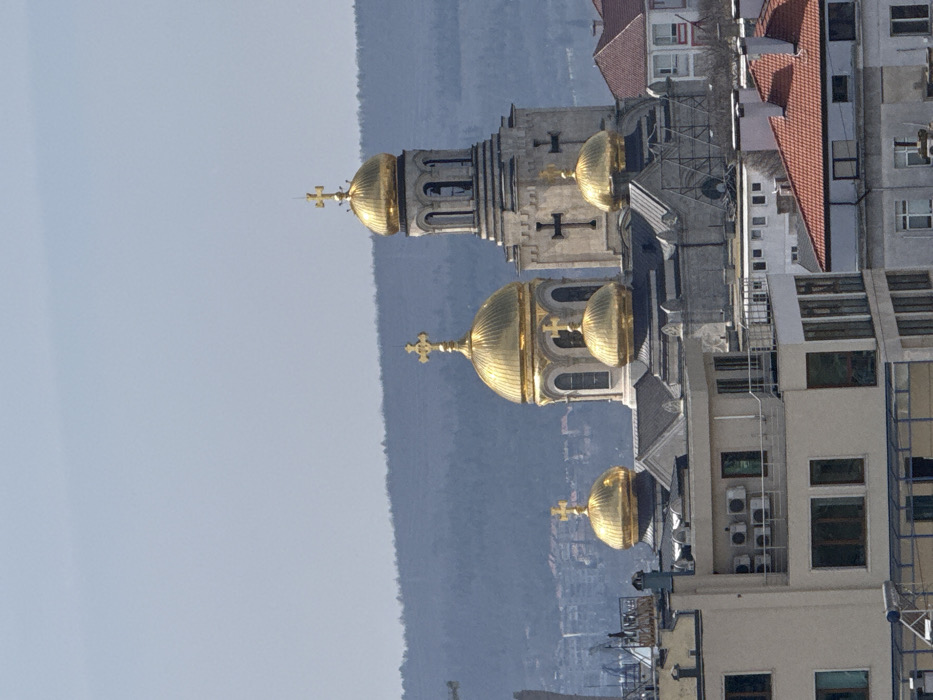 The image displays an elevated view of the Dormition of the Theotokos Cathedral in Varna, Bulgaria, situated in the city's central area. The cathedral, a prominent Orthodox church, features multiple golden domes topped with golden Orthodox crosses. Its main structure comprises a large central golden dome, a tall bell tower with its own golden dome and cross positioned to the right, and four additional smaller golden domes visible on other sections of the church roof. The cathedral walls are constructed from light-colored stone, possibly pale grey or white, and exhibit arched window openings and affixed black Orthodox crosses. Scaffolding is partially visible around the lower section of the cathedral structure.
In the foreground, several multi-story residential or commercial buildings with light-colored facades and various roof styles are present. One building on the left features multiple outdoor air conditioning units mounted on its exterior wall, along with a partially visible "PLANET" sign on its rooftop. Another building in the foreground has a sloped reddish-brown tiled roof. A section of a construction crane is visible on the far left edge of the frame.
The background consists of a clear, pale blue sky above a line of distant, tree-covered hills. Some sparse urban development is discernible at the base of these hills. No individuals or specific actions are identifiable in the scene. The lighting suggests daylight hours under clear weather conditions.