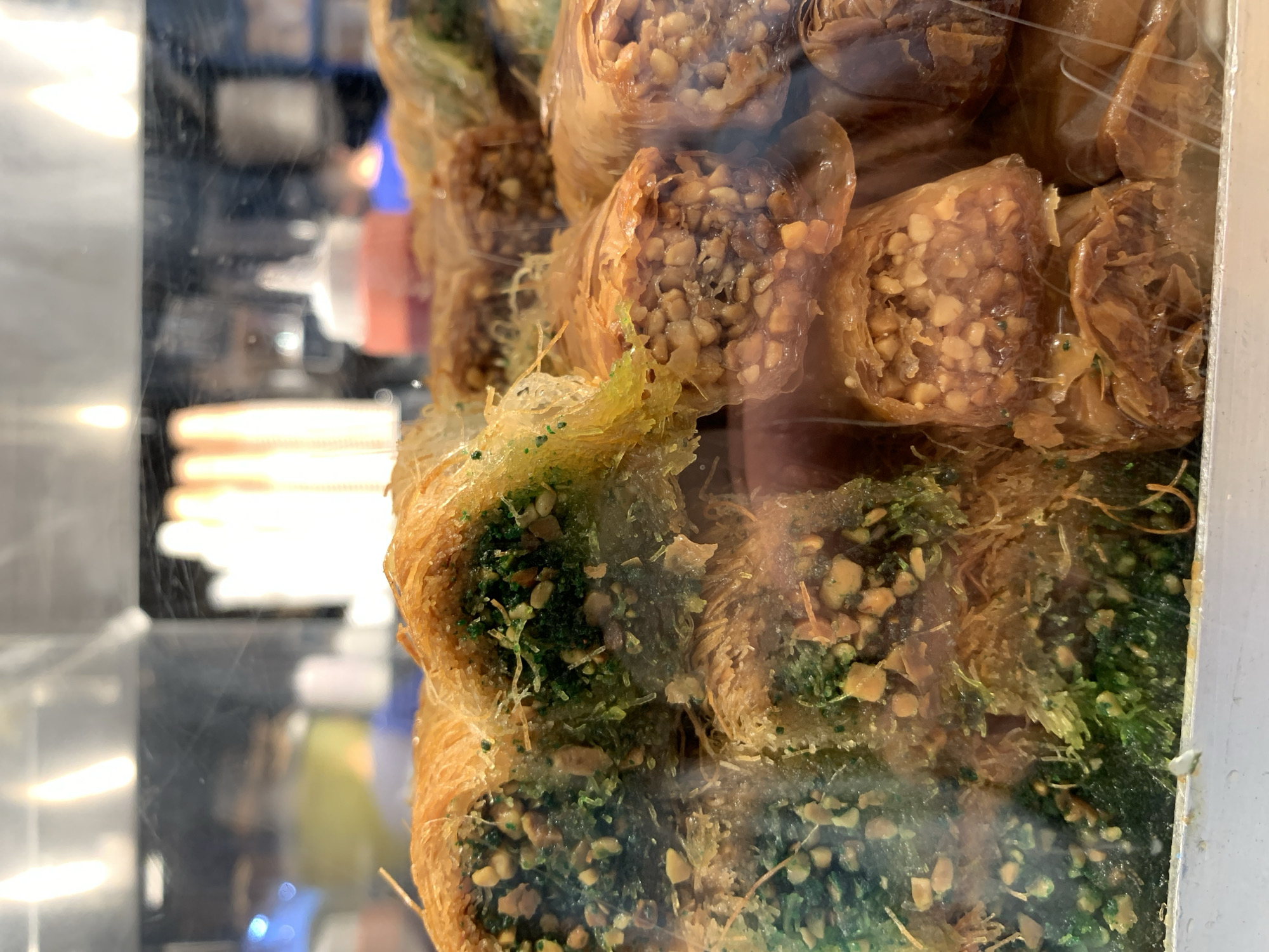 The image shows a close-up view of Middle Eastern pastries, specifically a type of rolled baklava, displayed in a glass case. Some are filled with nuts, while others are filled with green pistachio paste. The pastries are likely located in Tel-Aviv, Israel, based on the type of sweets shown which are commonly found in that city. The image is taken through the glass of a display case. The background is blurred, showing reflections of other objects and lights.
