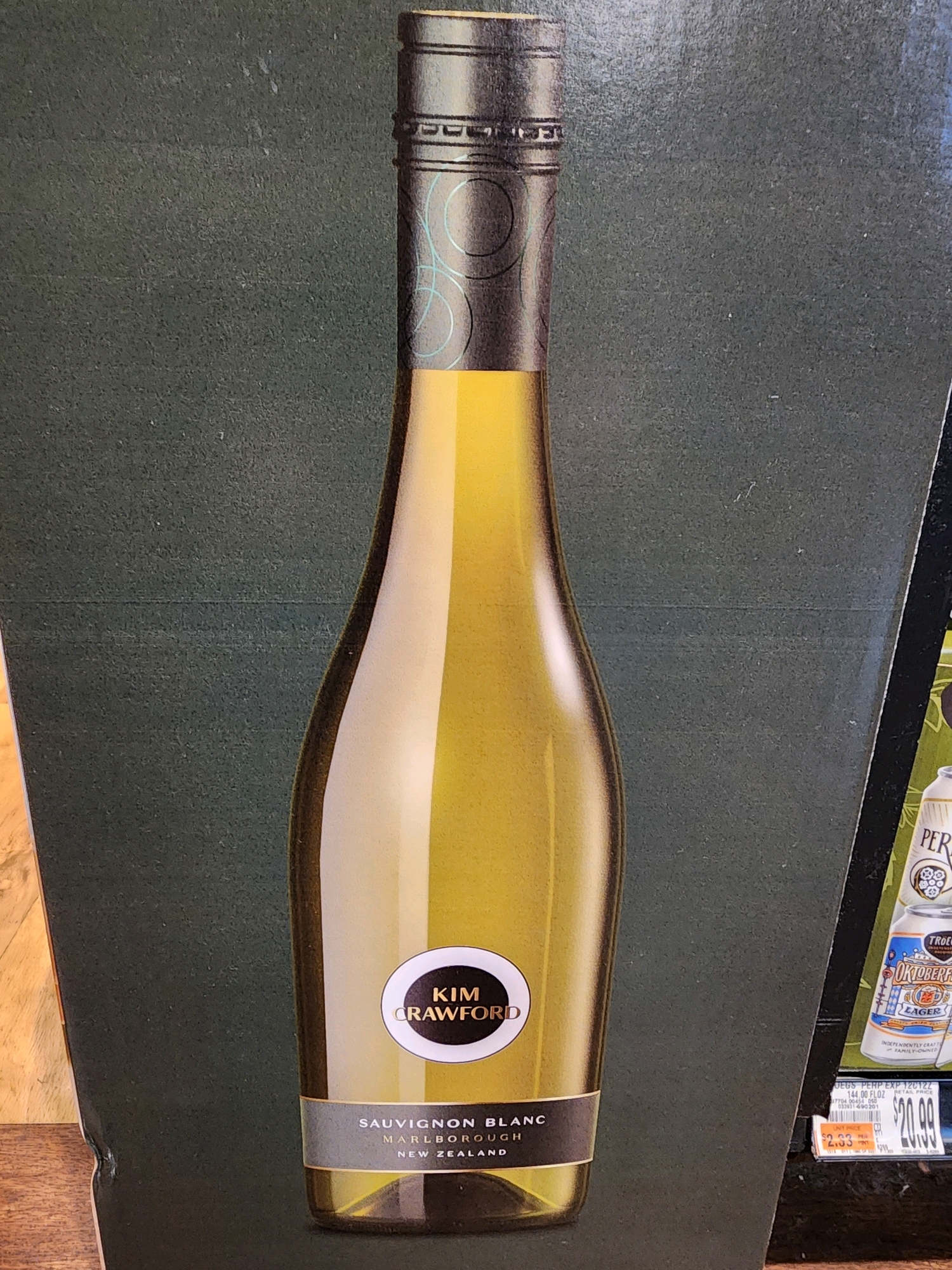 A full-frame, eye-level, horizontal, medium shot photograph displays a bottle of Kim Crawford Sauvignon Blanc wine. The bottle, rendered in yellow, stands against a dark green background. It has a metallic silver top and a label with a white circle containing the text "KIM CRAWFORD." Below the circle, a grey band with white text reads "SAUVIGNON BLANC MARLBOROUGH NEW ZEALAND." To the right of the wine bottle, a portion of a beer can labeled "Oktoberfest" is visible, with the text "TRÖEGS" and "LAGER" on it. Below the beer can, a price tag displays "$2.33" crossed out and "$20.99" below it, with the text "JEGS PERP EXP 12C122 144.00 FL OZ." The lighting in the scene is moderate, casting subtle reflections on the bottle. The overall impression is of a product advertisement or display.