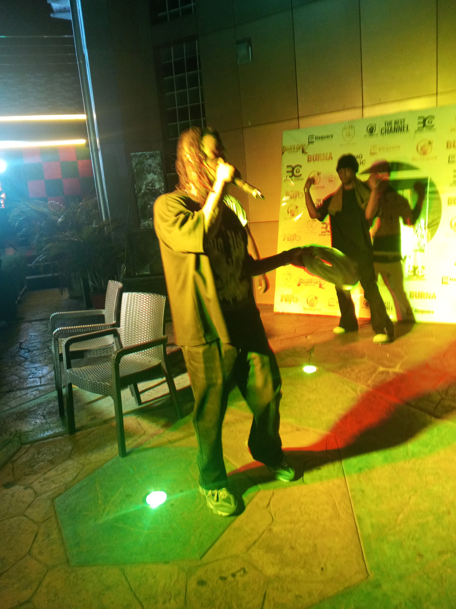Here is a description of the image:
The image shows a musician performing on a stage with two dancers behind him. The stage is lit with colorful lights, predominantly green and yellow. A backdrop behind the performers features logos and text. The main performer is holding a microphone and seems to be rapping or singing. To the side of the stage, there are three chairs. The location appears to be an outdoor venue, possibly in Lokoja, Nigeria, as indicated by the logos on the backdrop.