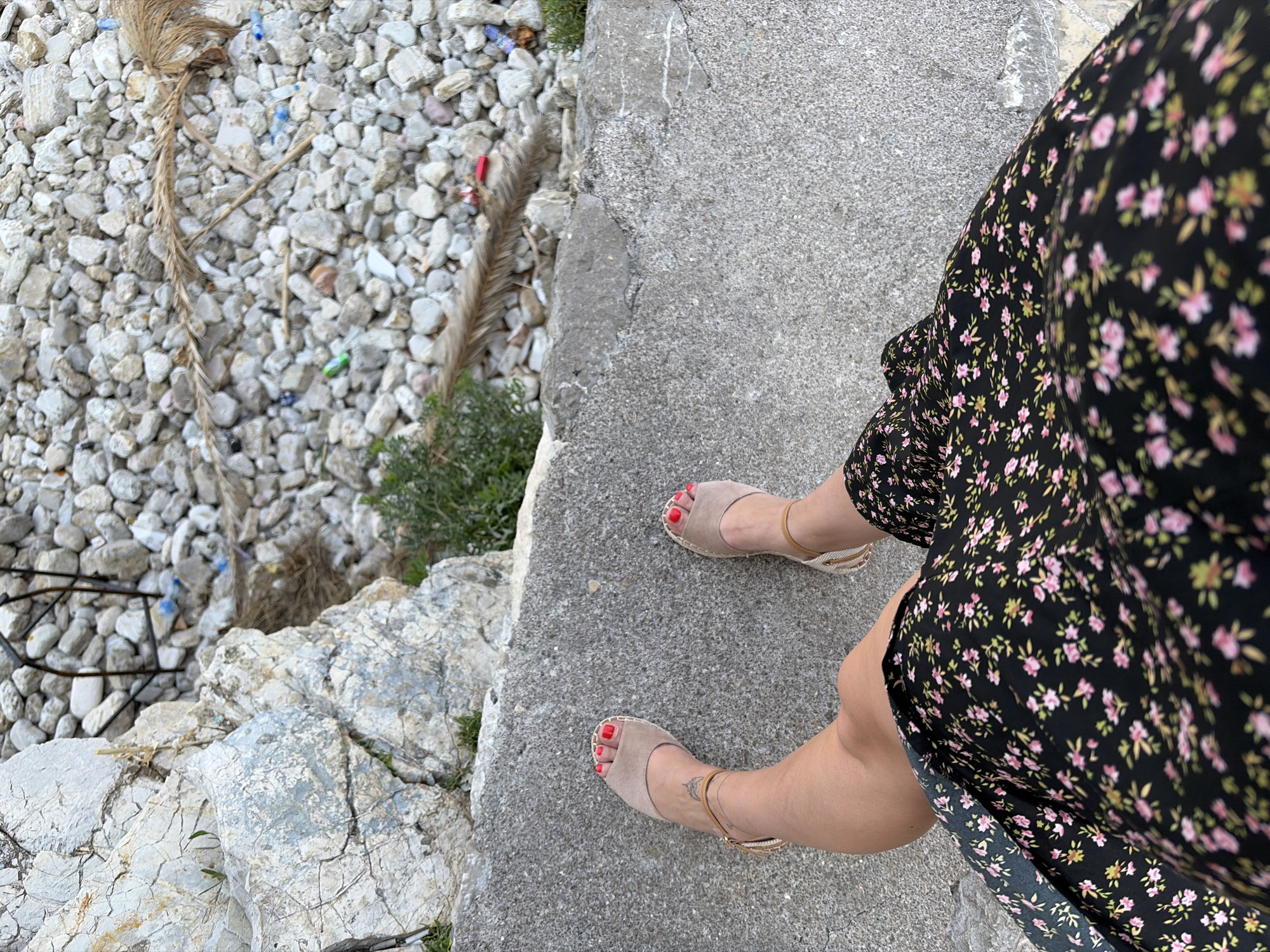 A low-angle shot shows a person's legs and feet from the knees down, standing on a grey concrete surface. The person is wearing a black floral print dress with small pink and green flowers, and taupe espadrille sandals with ankle straps. Red nail polish is visible on the toenails. To the left of the concrete surface is a large, rough, grey rock formation. Behind the concrete, the ground is covered in small, light-colored pebbles and some sparse green vegetation. Scattered amongst the pebbles are various pieces of debris, including plastic bottles and dried plant material. The overall scene suggests an outdoor setting, possibly a coastal area given the rocks and pebble ground. The lighting indicates it is daytime.