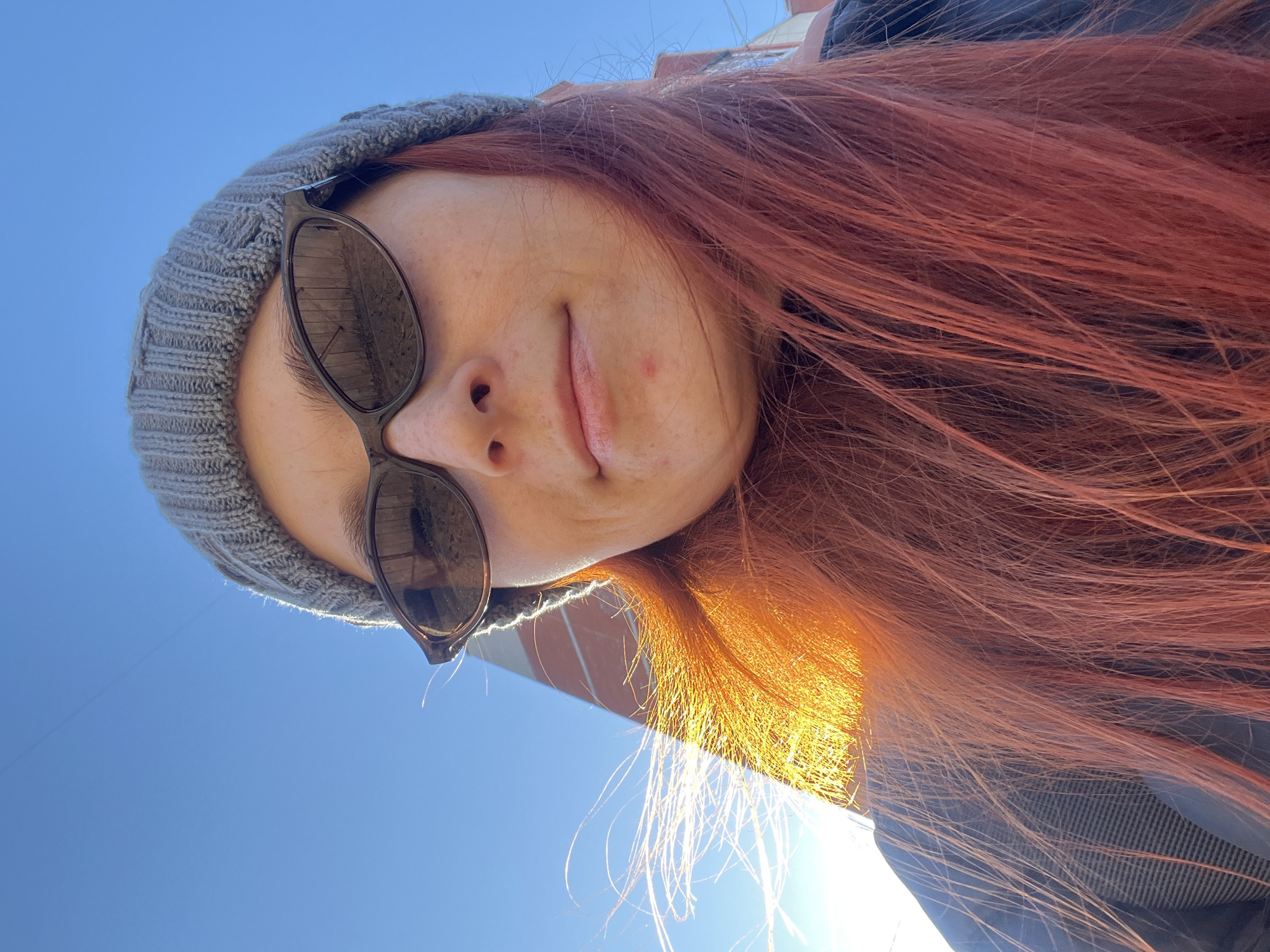 A woman with reddish-brown hair is wearing a grey knitted hat and sunglasses. Her face is tilted upwards, and she has a small blemish on her cheek. The background shows a clear blue sky and part of a brick building. The location is potentially Chaikovsky, Russia, as per the prompt.
