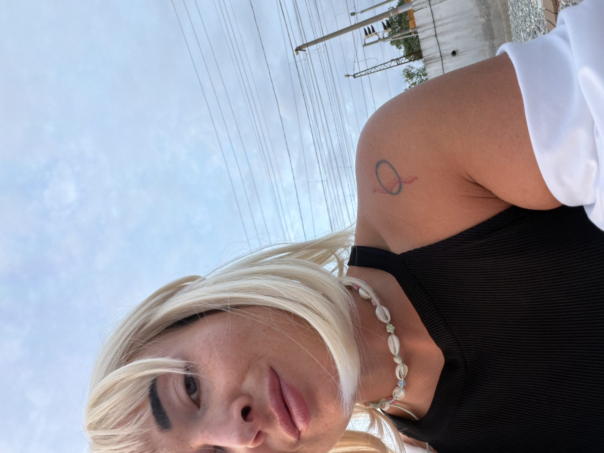 The image shows a close-up, waist-up shot of a woman with blonde hair and bangs, wearing a black tank top and a white jacket draped over her shoulders. She is also wearing a necklace made of shells and beads. A small tattoo is visible on her upper left arm. The background features a cloudy sky with overhead power lines and poles, suggesting an outdoor urban or industrial setting, possibly near a train track given the overhead wires and general aesthetic. The scene is lit by natural light. The woman is looking towards the camera with a slight smile. The location is specified as the city of Sochi, Russia.