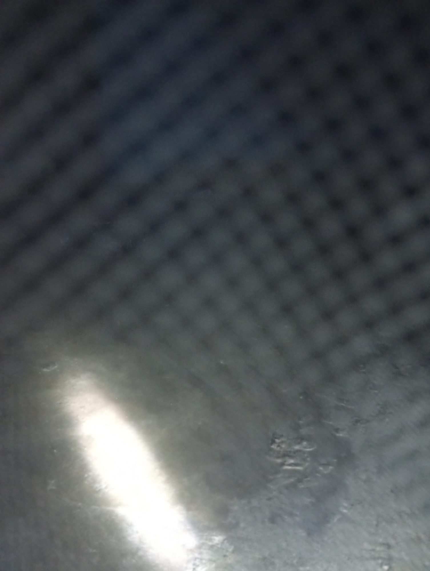 The image displays a textured, dark surface with a faint grid-like pattern visible throughout. A bright, elongated reflection streaks across the lower left portion of the surface. In the lower right area, there are subtle, embossed markings that are difficult to decipher due to blurriness. The overall impression is of a reflective, possibly metallic or plastic material with some form of indentation or texture. There are no discernible people, actions, or specific environmental features that would allow for identification of a location within Jalingo, Nigeria.