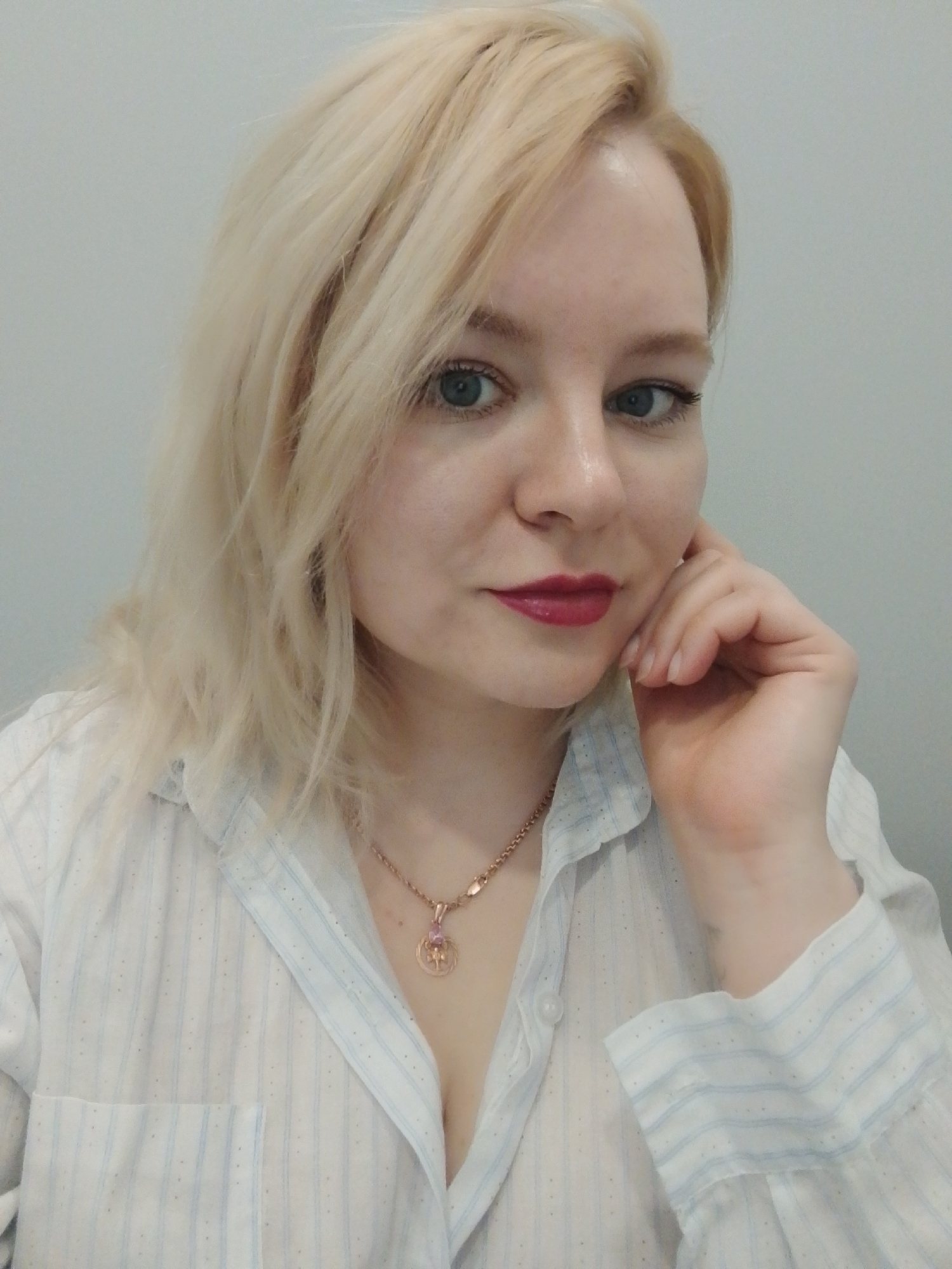 A medium shot depicts a fair-skinned woman with blonde, shoulder-length hair. Her hair is styled with loose waves. She has light blue eyes and her lips are painted with a dark red lipstick. She is wearing a white button-down shirt with light blue vertical stripes. A gold necklace with a circular pendant is visible around her neck. Her right hand is raised to her face, with her fingers lightly touching her cheek. The background is a plain light gray wall. No information related to Izhevsk, Russia can be discerned from the image.