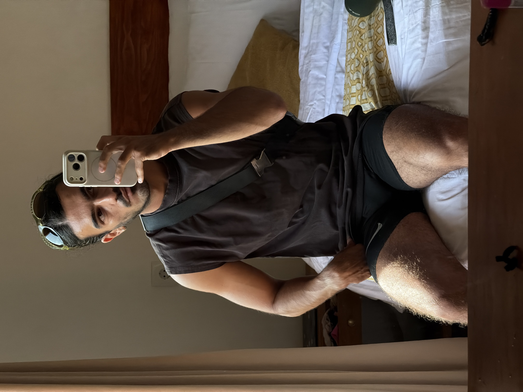 The image is a mirror selfie showing a young man with dark hair, a beard, and sunglasses perched on his head. He is wearing a dark, short-sleeved t-shirt and black athletic shorts, which have a white Nike swoosh visible on the left thigh. A dark strap, likely from a bag or a camera, crosses his chest. He holds a light-colored smartphone, possibly an iPhone with a triple-lens camera, in his right hand to capture his reflection.
The setting appears to be an indoor space, likely a bedroom or hotel room, reflected in the mirror. Behind the man, a bed is visible with white sheets, a mustard yellow pillow, and a white and gold patterned throw blanket. A dark wooden headboard is also discernible. The room features light-colored walls, and an electrical outlet can be seen on the wall in the upper background (if the image were oriented vertically). The man's legs are visibly hairy, and his left hand rests on his thigh. The image itself is rotated 90 degrees counter-clockwise, making the reflected man appear to be lying down horizontally. The only visible text is the Nike swoosh on his shorts. The location context is North Kuta, Indonesia.