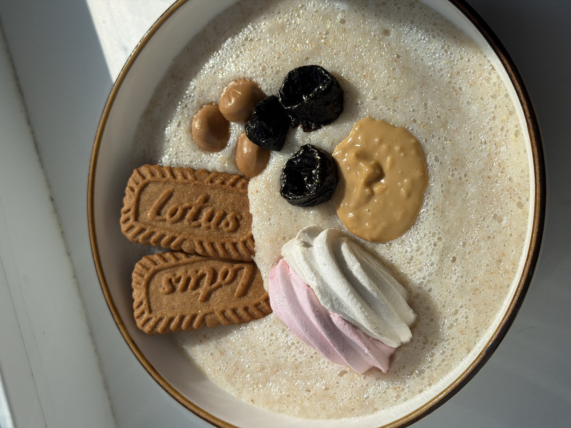 A top-down shot shows a light brown bowl filled with a frothy, light-colored liquid, possibly a smoothie or milk-based beverage. Scattered across the surface of the liquid are several toppings. To the left, two rectangular biscuits with ridged edges are partially submerged. The top biscuit has "Lotus" embossed on it, and the bottom biscuit has "Biscoff" embossed.
Clustered towards the center of the bowl are four dark, dried fruits, likely prunes, and three small, dollop-sized mounds of a light brown paste, possibly peanut butter or a similar spread. To the right of these, a larger, flatter dollop of the same light brown paste is visible.
At the bottom center of the bowl, a decorative swirl of meringue is placed, with the top portion being white and the bottom portion being pink. The lighting suggests it is daytime, with shadows cast from the left, indicating sunlight. The setting appears to be indoors, likely a kitchen or dining area, with a light-colored surface visible around the bowl. No people are visible. The overall impression is of a breakfast or snack item, with sweet and creamy elements.