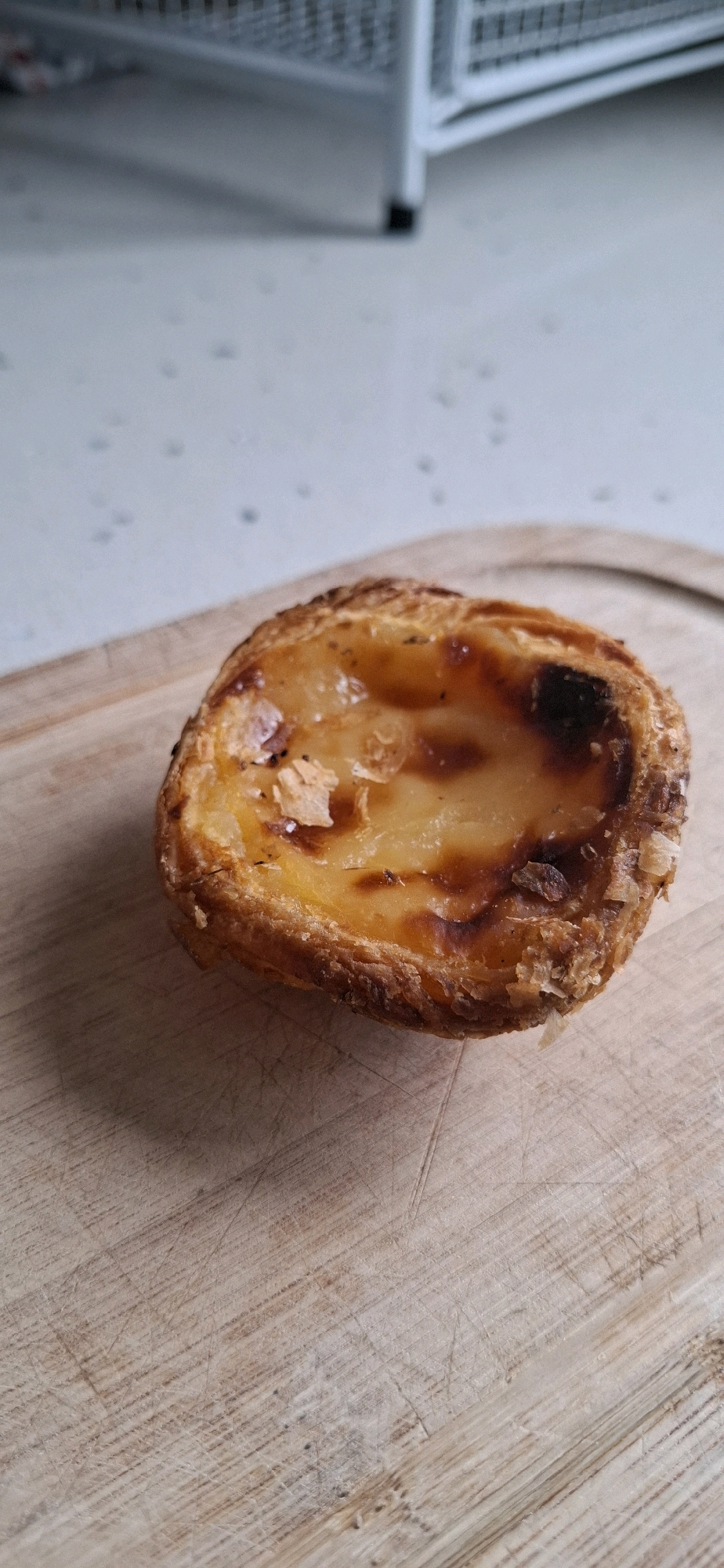 A single Portuguese egg tart, a pastel de nata, rests on a wooden cutting board. The tart has a flaky, golden-brown crust and a creamy, caramelized custard filling that is slightly browned in spots. The cutting board is light-colored wood with visible scratch marks. The background is out of focus, showing a light-colored textured surface and the edge of a white shelving unit. There are no people or any indication of a specific city or location.