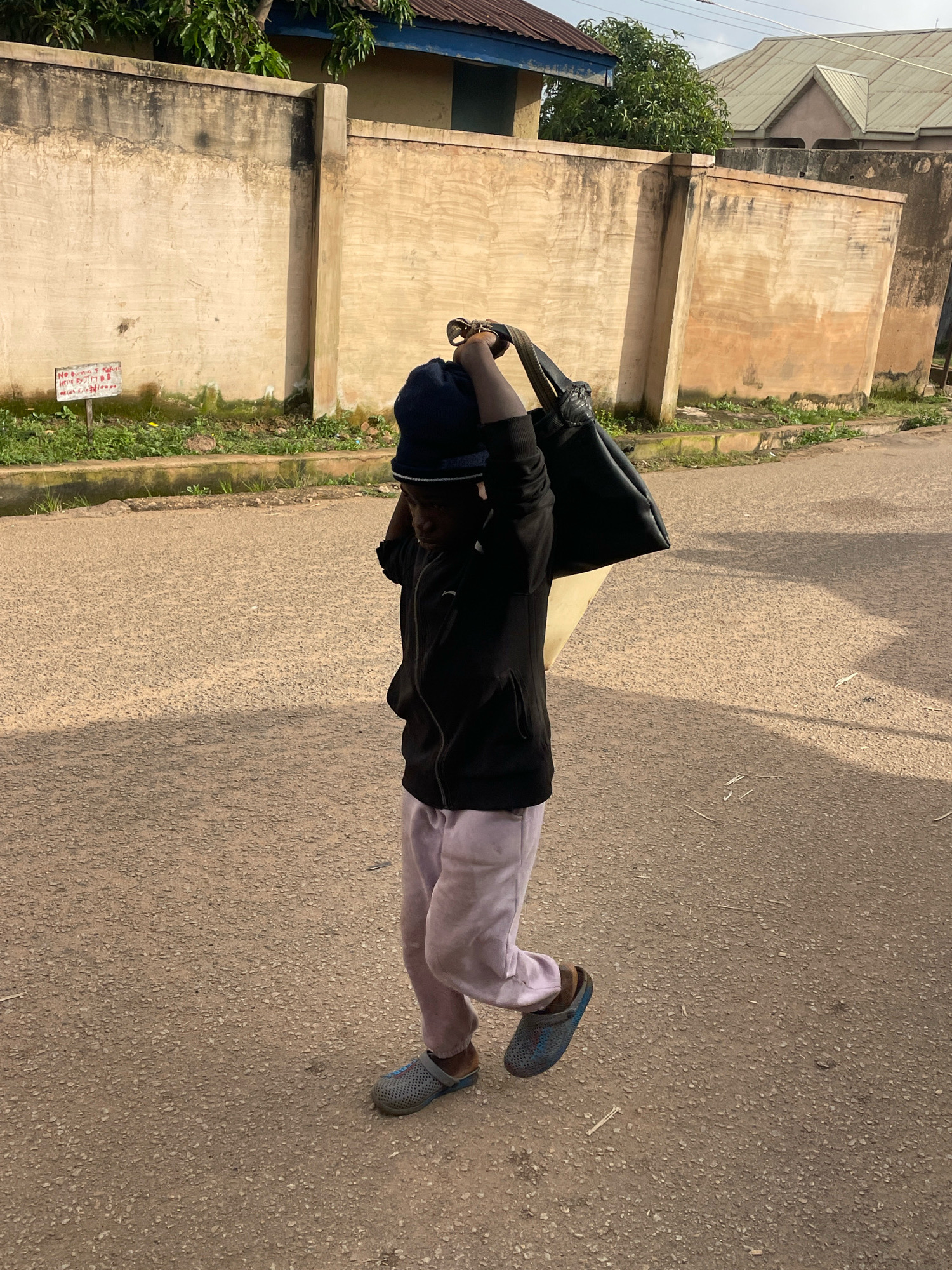 A full-body, eye-level shot captures a person from the waist up, walking on a paved street in Jos, Nigeria. The person, wearing a dark blue beanie, black hooded jacket, light purple pants, and blue clogs, carries a large black bag over their right shoulder. Their head is bowed, looking down. The street's surface is rough and asphalted. Behind the person, a beige wall, stained with dark marks, runs horizontally. A small white sign with red lettering is affixed to the wall. Green vegetation grows along the base of the wall, and a curb is visible in the foreground. In the background, houses with pitched roofs are discernible. The sun casts a shadow of the person onto the street.