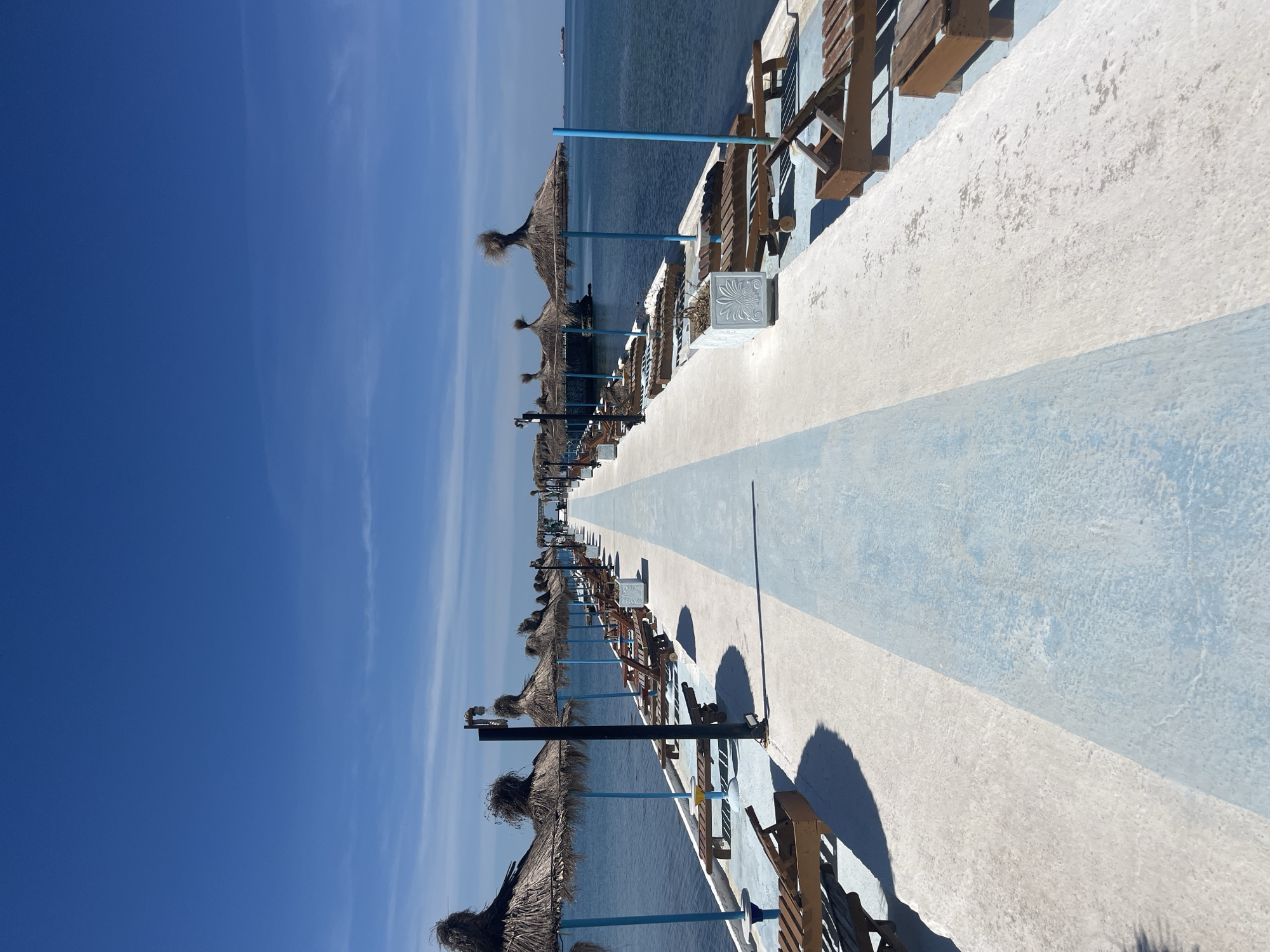 Here is a description of the image:
This image shows a waterfront pier or walkway, seemingly in the city of Unknown, Unknown. The pier is marked with a light blue and white painted pathway. Along one side of the pier, there are multiple beach umbrellas, constructed of straw or similar natural material, placed to provide shade. There are also wooden lounge chairs and a low wall lining the edge of the pier, offering seating and structure. Beyond the pier, there's a view of the water under a clear blue sky with some light cloud cover. A large ship can be seen in the distance on the water. The lighting suggests it is a sunny day.