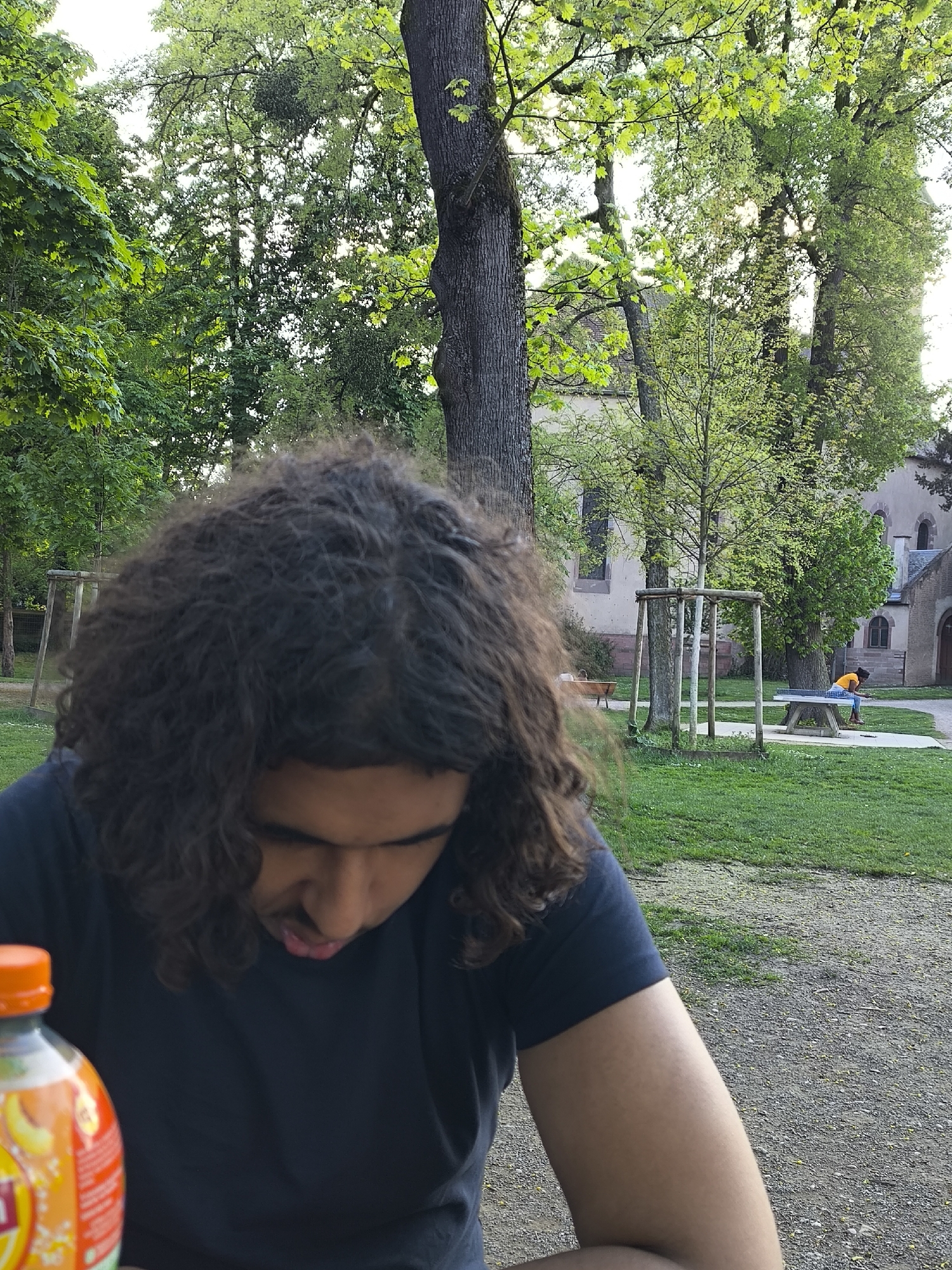 The image shows a man with curly hair, wearing a dark t-shirt, seated outdoors. In the background, there's a grassy park with trees and a building that appears to be part of the Saint-Pierre-le-Jeune Protestant church in Strasbourg, France. A person in an orange shirt is sitting at a ping pong table in the background. A bottle with an orange cap sits on a table in the foreground. The man is looking downwards.