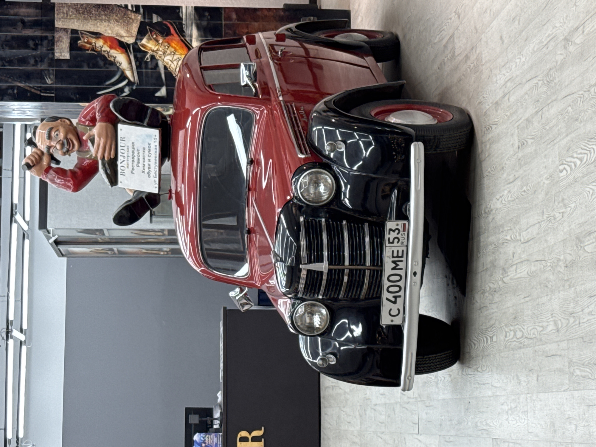 The image features a vintage car with the license plate "C 400 ME 53 RUS". Adjacent to the car is a statue of a mustachioed man holding a hammer and a sign that reads "BONJOUR мастерская Реставрация Ремонт Химчистка обуви и сумок ул. Бестужевская 10". The location appears to be the city of Saint Petersburg, Russia. The scene includes elements of automobile restoration and potentially shoe repair, suggesting it is a workshop or related commercial establishment.
