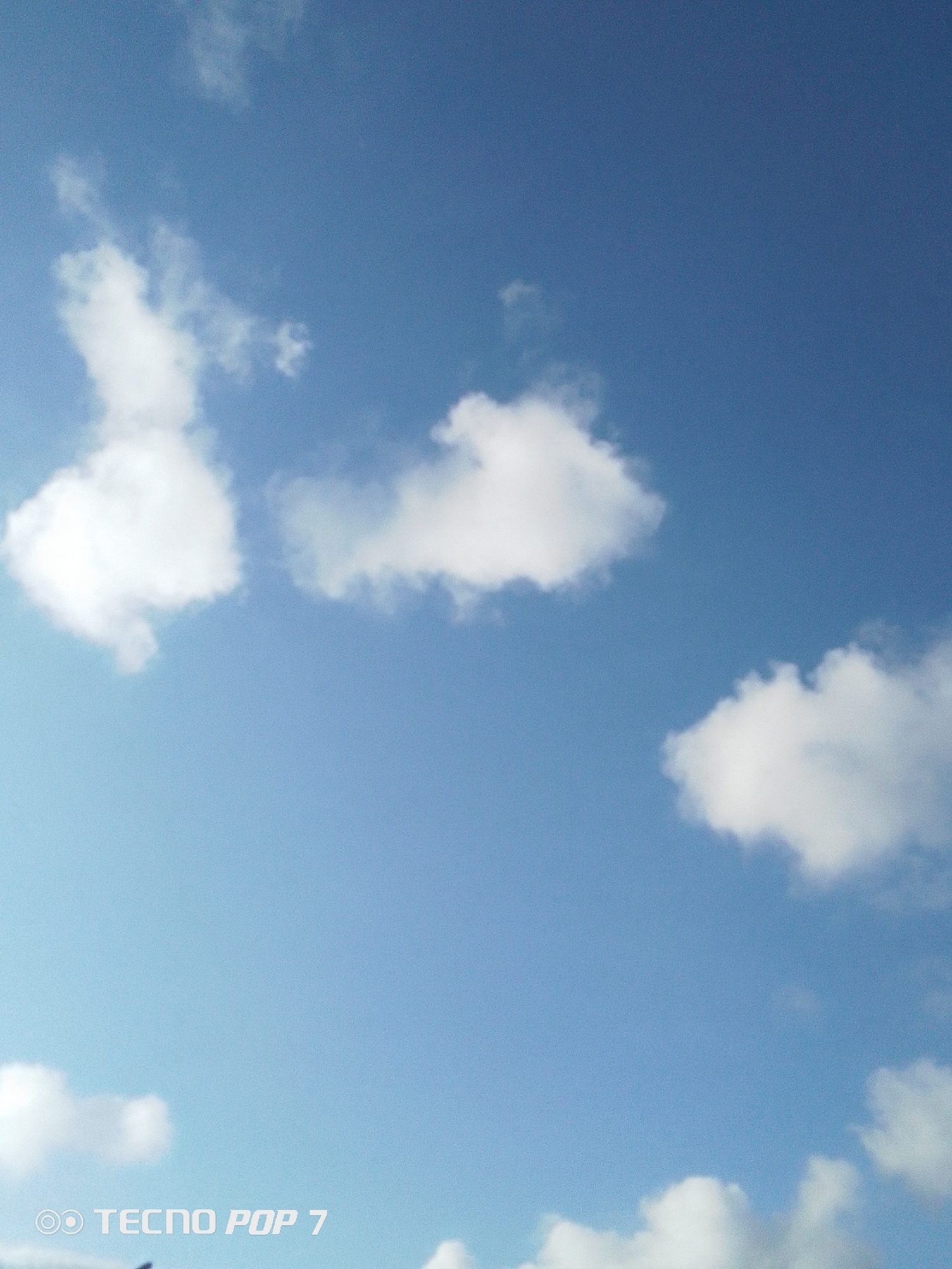 The image shows a daytime sky with scattered white clouds. The sky is a clear blue. The photograph was taken in Pedro González, Venezuela, as evidenced by metadata not present in the image. There are no people or objects other than clouds visible. The TECNO POP 7 watermark is visible in the bottom left corner.