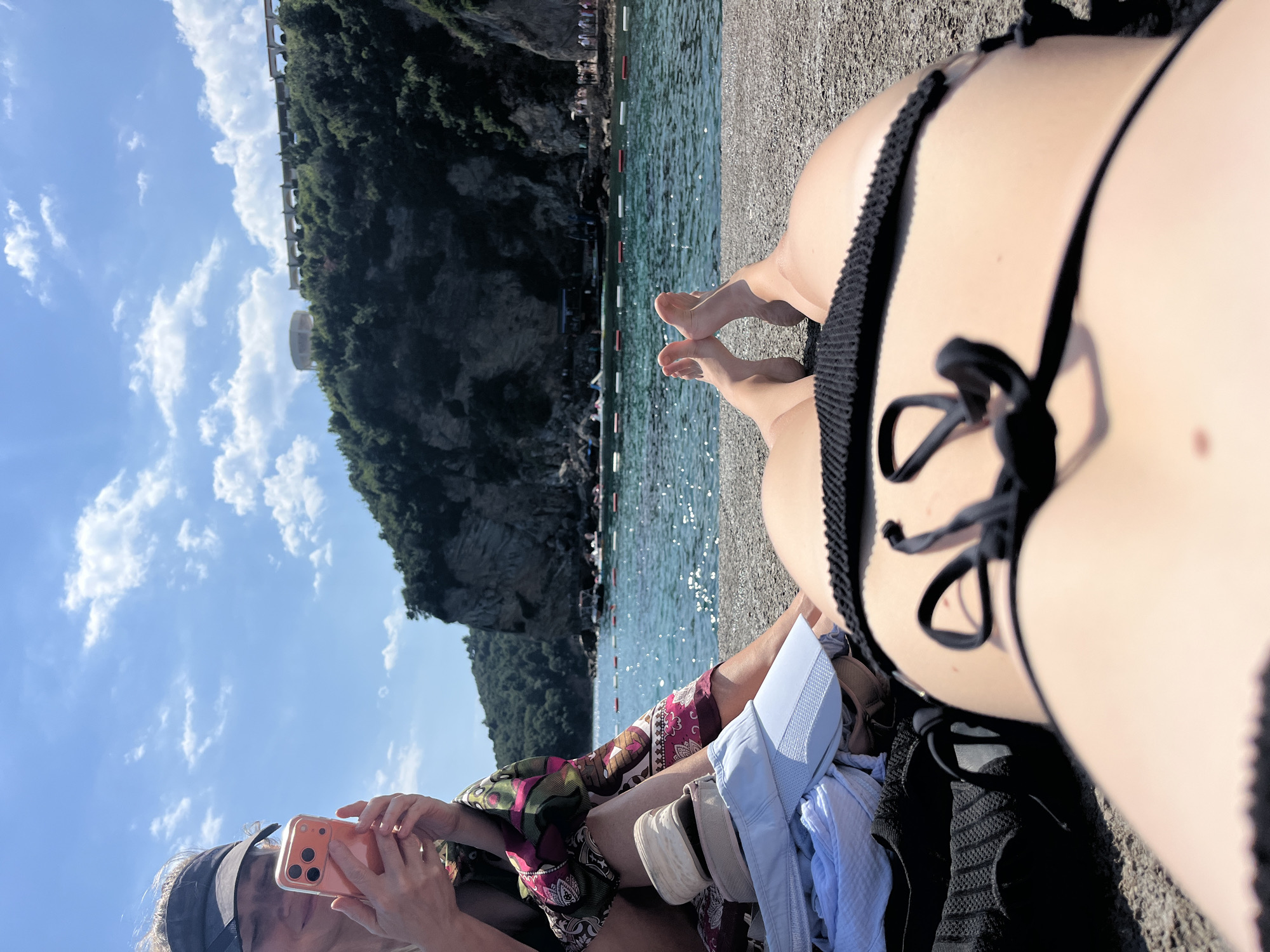 The image captures a sunny day at a beach in Budva, Montenegro. The foreground is dominated by the lower body of a person wearing a black bikini. Next to them, another person is partially visible, holding a peach-colored smartphone. They are dressed in casual beach attire, including a patterned sarong and a light blue shirt. Scattered around them are various beach items like a hat and a black knitted sweater.
In the background, a picturesque coastline unfolds. The turquoise waters of the Adriatic Sea are calm, with a thin line of buoys marking a swimming area. Beyond the water, lush green hills rise steeply, dotted with trees. A walkway or road can be seen carved into the hillside, with some buildings or structures visible. The sky is a bright blue, with scattered white clouds, indicating clear and pleasant weather. The overall impression is one of relaxation and enjoyment on a beautiful summer day. The image appears to be taken from a low angle, looking up towards the sky and the scenic background. No visible text is present in the image.