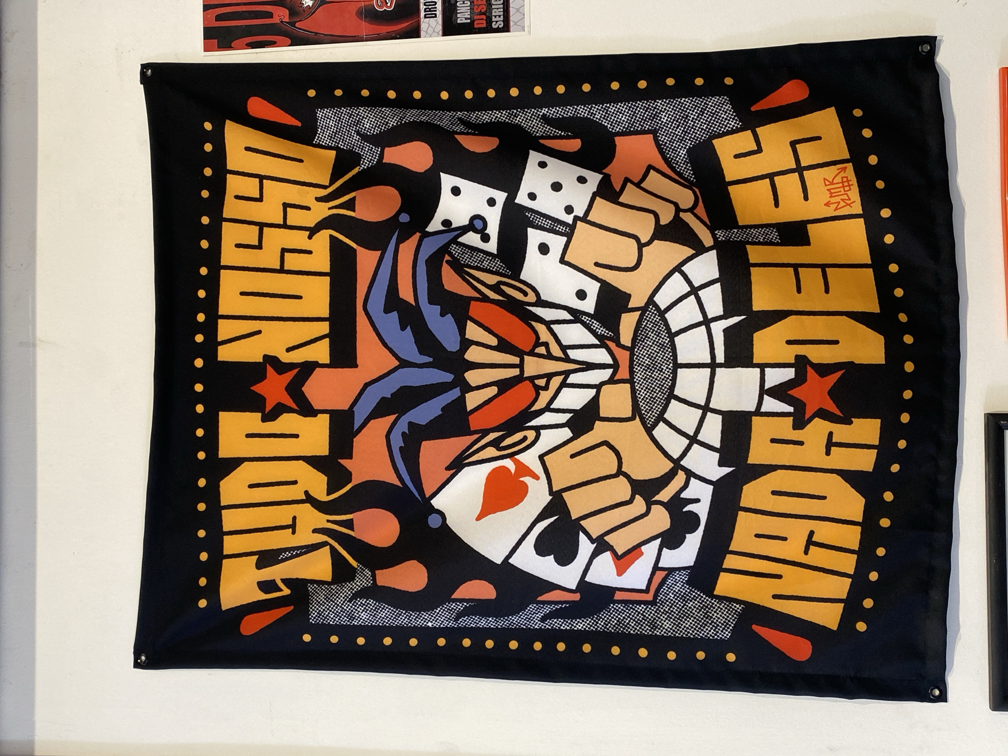 A vibrantly colored fabric banner, oriented horizontally, hangs against a plain white wall. The banner features a central graphic design with a bold black border punctuated by small yellow-gold circular dots.
The design's focal point is a stylized jester or clown head, predominantly blue, red, and black, positioned centrally and vertically inverted. Flanking the jester are human-like hands rendered in a light skin tone with black outlines. These hands are depicted holding gambling elements: white dice with black dots and playing cards featuring a red heart and a black spade. The background of the central graphic includes abstract, flame-like shapes in orange and black, along with halftone-style dotted patterns and a grid-like design in white and black.
Large, blocky, yellow-gold text is integrated into the design. The word "WHOSE" is spelled across the top half of the banner, inverted, while "SIDE" is spelled across the bottom half, also inverted. "ARE" runs vertically along the left side, and "YOU" runs vertically along the right side. On the 'E' of "ARE," there is a small, stylized red signature or text that appears to read "2R2K" or similar characters.
The banner is secured to the wall by metal grommets in its top corners and potentially in the bottom corners as well, though only one is clearly visible. Above and to the right of the banner, a portion of another graphic or poster is visible, featuring red and black colors and partial text like "DROP," "PANCA," and "DJ SE." The overall imagery on the main banner suggests themes of chance, games, and identity, encapsulated by the direct question "WHOSE SIDE ARE YOU." The setting is an indoor space, possibly a gallery, studio, or retail environment in Buenos Aires, Argentina.