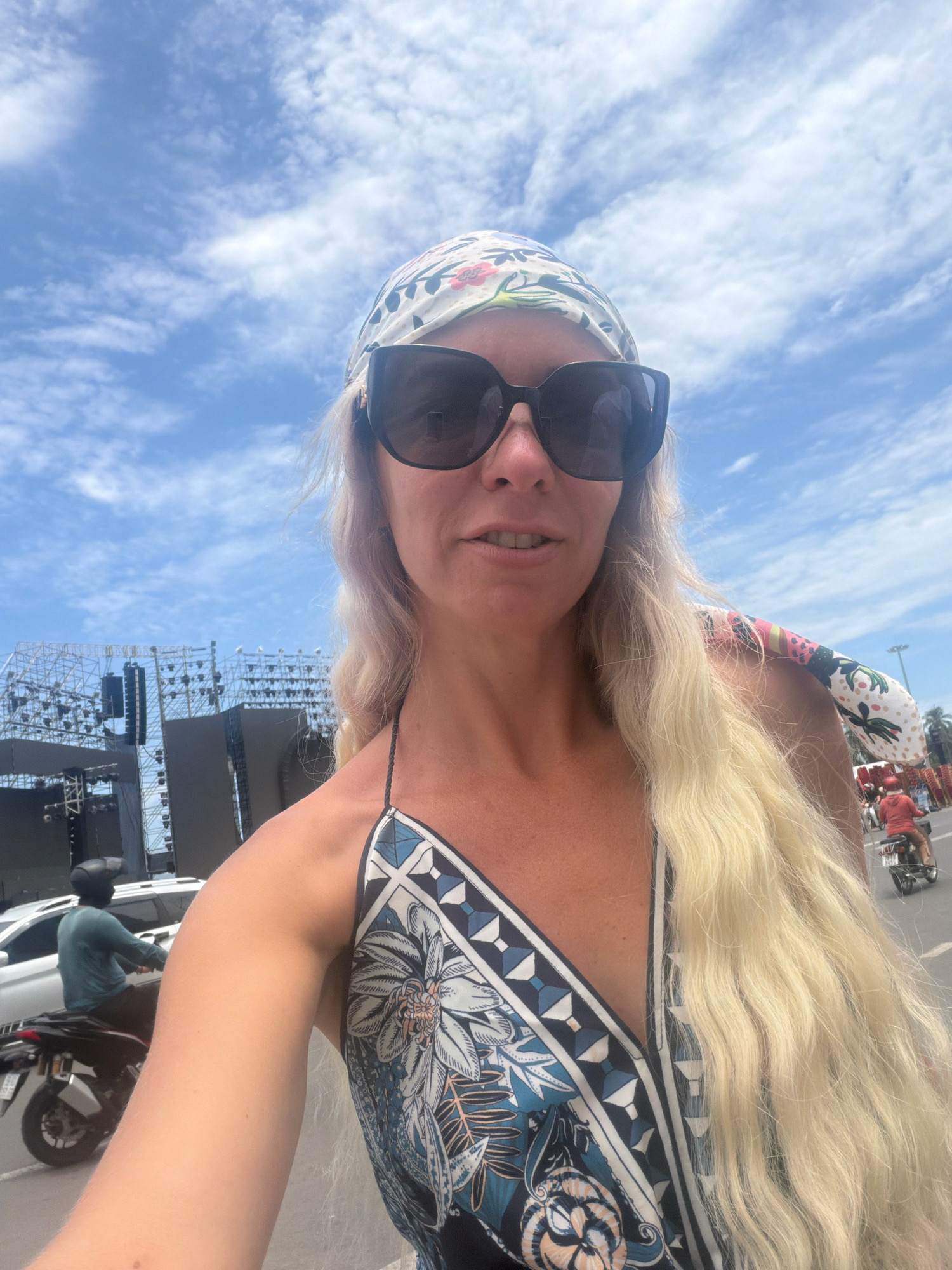 A woman with long blonde hair, wearing large dark sunglasses and a floral bandana, takes a selfie on a street in Nha Trang, Vietnam. She is wearing a patterned dress. In the background, a large stage setup with scaffolding and speakers is visible, along with a white car and several motorbikes carrying people. The sky is blue with scattered clouds.