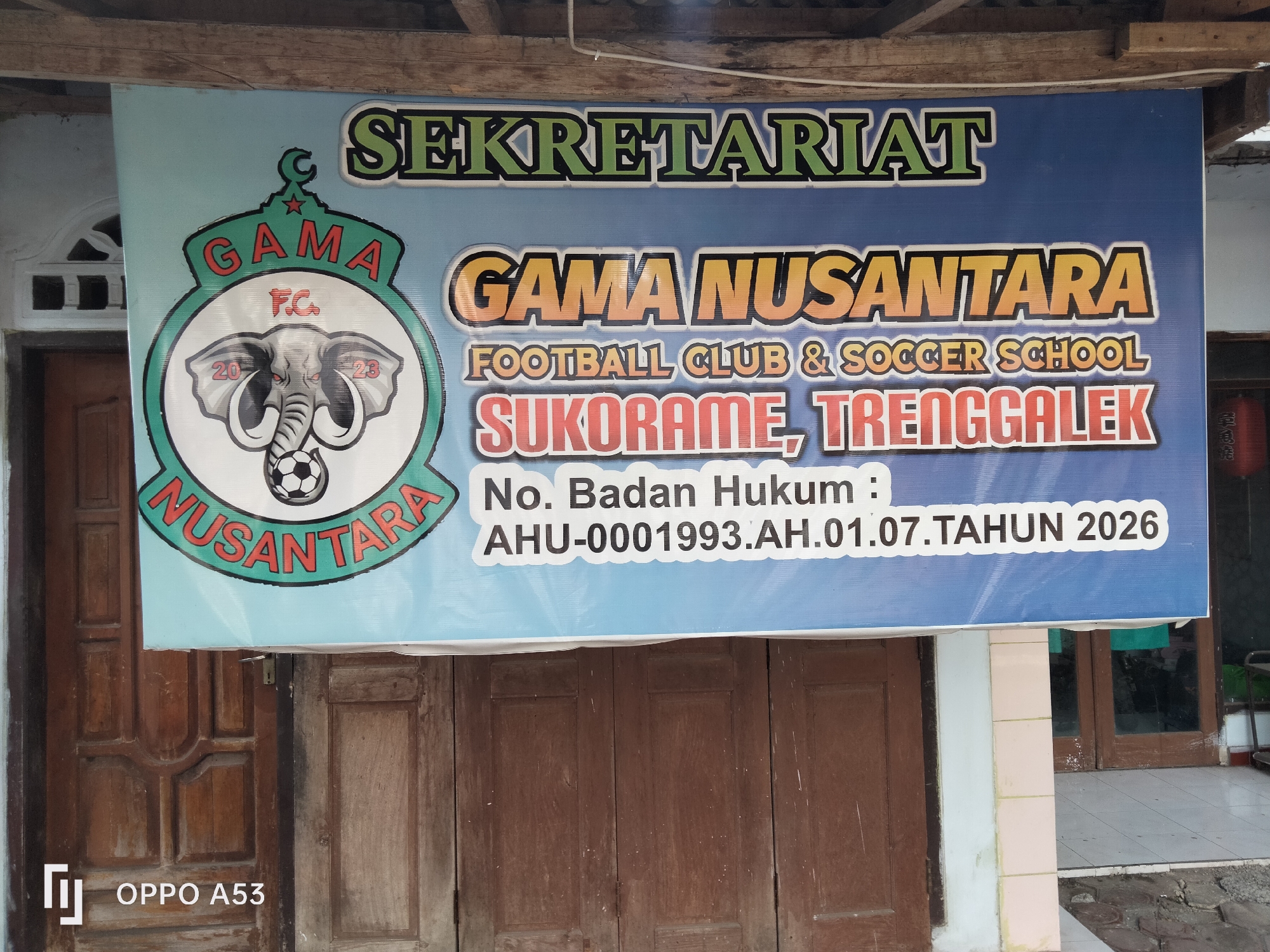The image shows a banner hanging outside a building. The banner has blue and light blue colors and features text and a logo. The text on the banner includes "SEKRETARIAT", "GAMA NUSANTARA", "FOOTBALL CLUB & SOCCER SCHOOL", "SUKORAME, TRENGGALEK", and "No. Badan Hukum : AHU-0001993.AH.01.07.TAHUN 2026". The logo on the left side of the banner is a circular design with an elephant's head holding a soccer ball. Below the banner, there are two wooden doors. The building appears to be made of wood and has a tiled floor in front. The overall scene suggests an outdoor setting in front of a facility related to football and soccer.