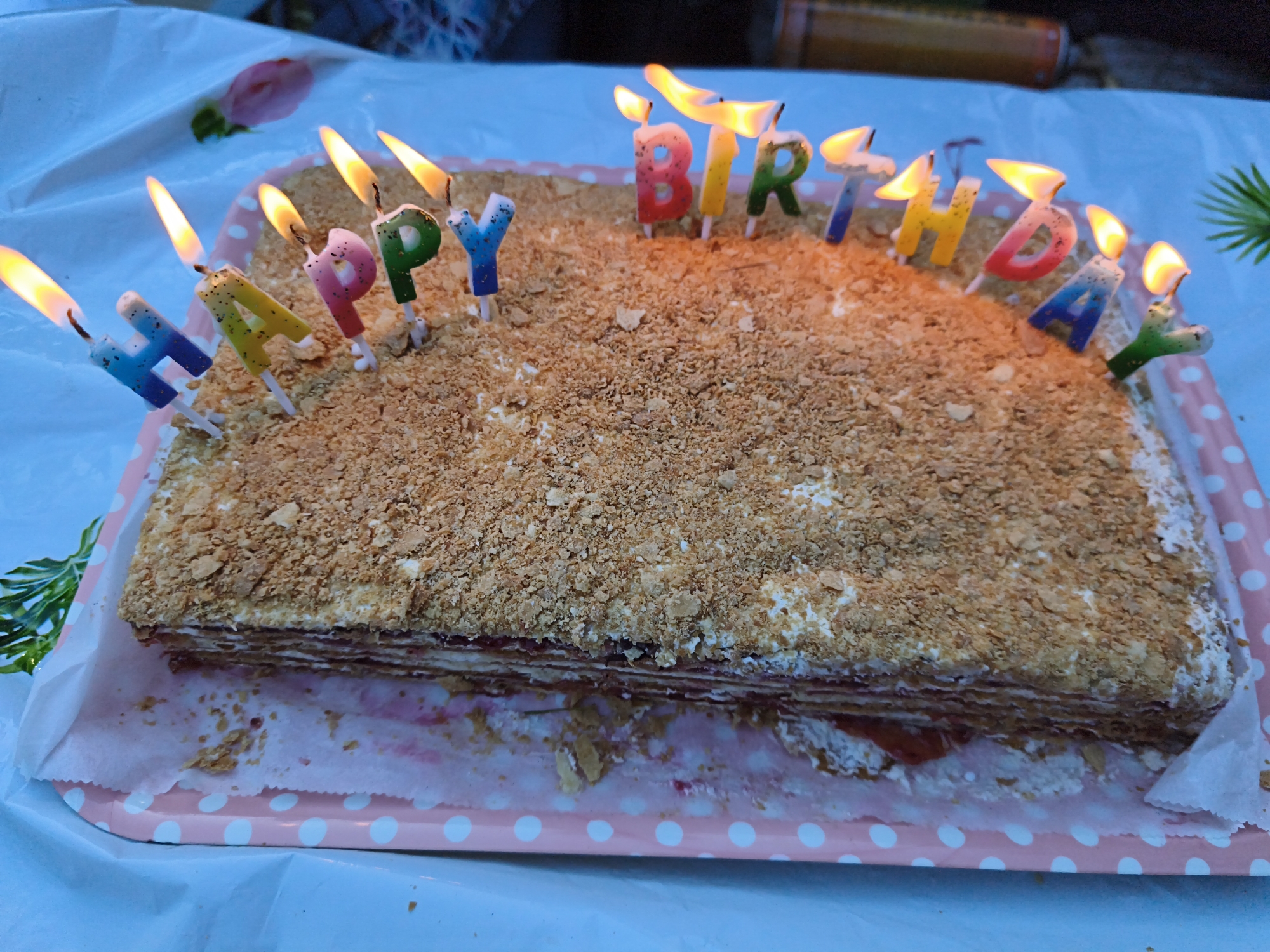 A rectangular cake, likely a layered Napoleon cake, is placed on a pink and white polka-dotted serving tray. The cake is covered in crushed nuts or pastry crumbs, with some white frosting visible. Numerous colorful birthday candles, lit and in the shape of letters, spell out "HAPPY BIRTHDAY" across the top of the cake. The candles are blue, green, pink, and yellow, with flames flickering. The cake and tray are set on a light blue surface, possibly a table covered with a tablecloth, with some greenery and a blurry object in the background. The scene appears to be outdoors, suggested by the lighting and the presence of foliage. There are no people visible in the image.