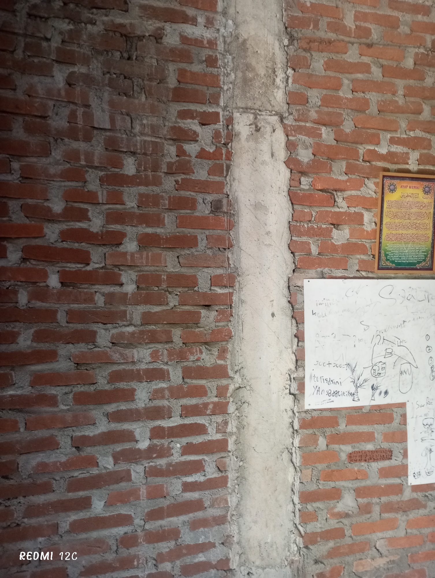 The image presents a section of a brick wall in Lhokseumawe, Indonesia. The wall is bisected by a vertical, roughly finished concrete column. To the left of the column, the brickwork is exposed. To the right, a framed decorative item displaying Arabic text hangs on the wall, next to a whiteboard with writing and drawings. The bottom left corner of the image displays the text "REDMI 12C," likely indicating the type of camera used to capture the image. The scene is focused on the physical structure of the wall and the items affixed to it.