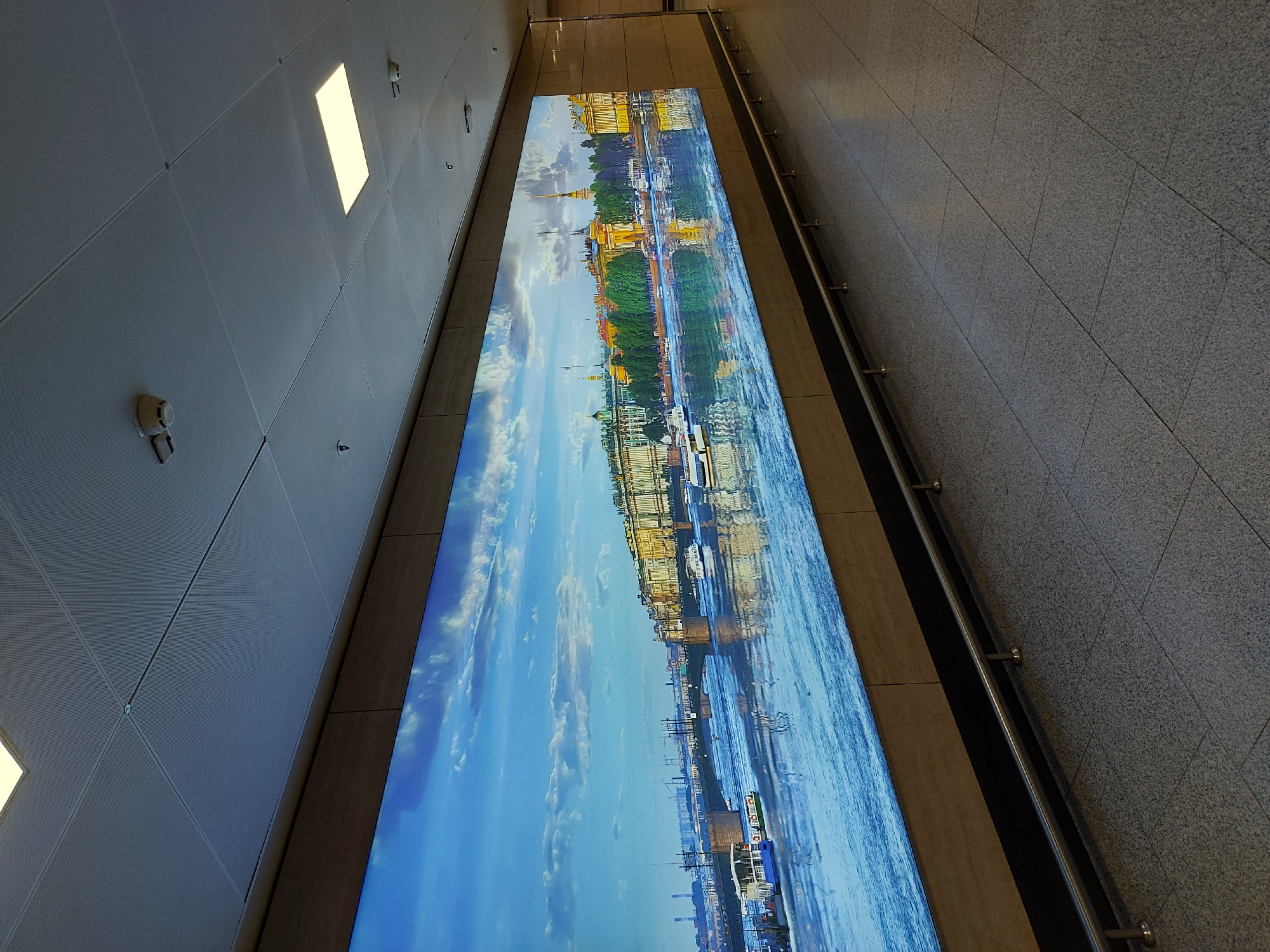 The image shows an indoor setting, likely a subway station or underground walkway, featuring a large, horizontally oriented digital display along one wall. The display features a scenic view of Saint Petersburg, Russia, showing the Neva River with historic buildings along its banks. The scene appears to be a reflection of the city's architecture and waterways, with a clear blue sky and clouds. The display is integrated into the wall, which is lined with stone-like tiles. A metal handrail runs alongside the display. The ceiling is made of panels with recessed lighting. The overall environment appears modern and clean. No people are visible in the image.