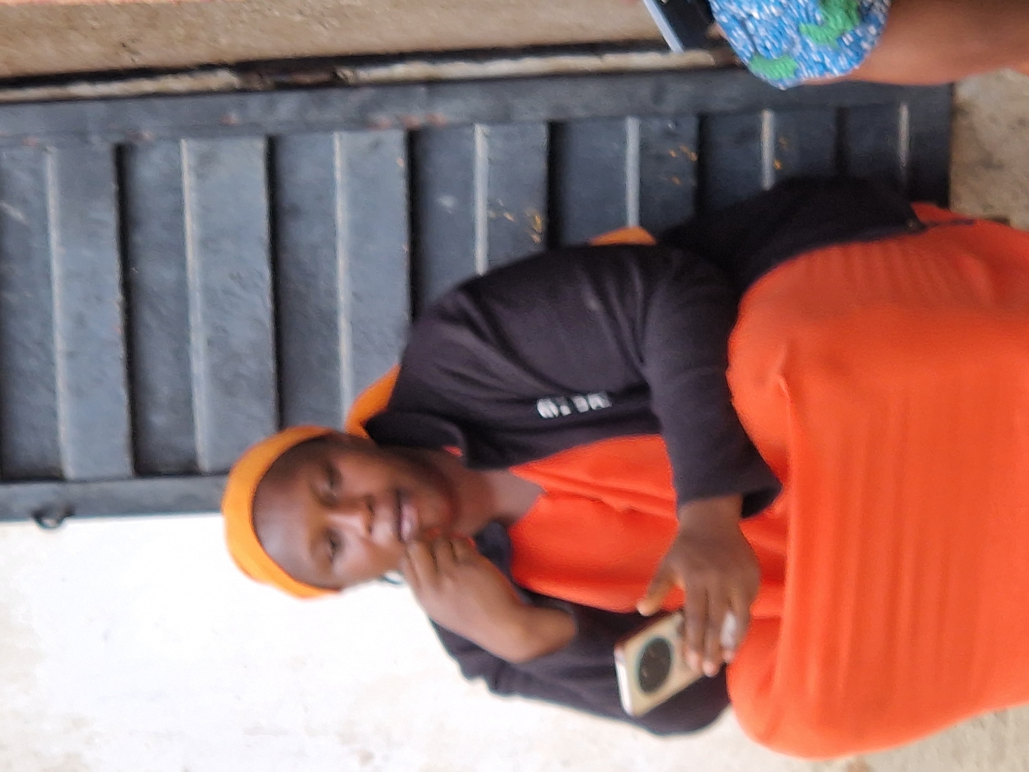 The image shows a woman wearing an orange headscarf and an orange dress, with a dark jacket over it. She is holding a smartphone in her left hand and her right hand is near her mouth. She is sitting in front of a dark, slatted structure, possibly a door or shutters. The background is out of focus, with a textured wall to the left and a hint of another person in patterned clothing on the right. The scene appears to be outdoors in a somewhat rustic urban environment, consistent with a location in Kabong, Nigeria.