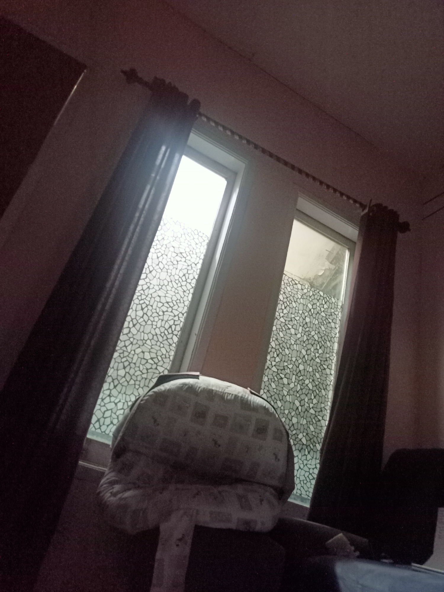 The image depicts an indoor scene, likely a residential room, with two windows side-by-side. The windows are adorned with a decorative film featuring a mosaic-like pattern of irregular shapes in shades of grey and white. Dark, heavy curtains are partially drawn on either side of the windows, obscuring the view of the outside. In the foreground, a large, plush object, possibly a bolster pillow or cushion, rests on a piece of furniture, its patterned surface visible. The overall lighting is dim, suggesting it might be nighttime or a heavily shaded area, with some light filtering through the windows. The room appears to be tilted in the photograph. There is no discernible activity or interaction taking place within the frame. The image does not provide any visual cues to indicate its location within Randuagung, Indonesia.