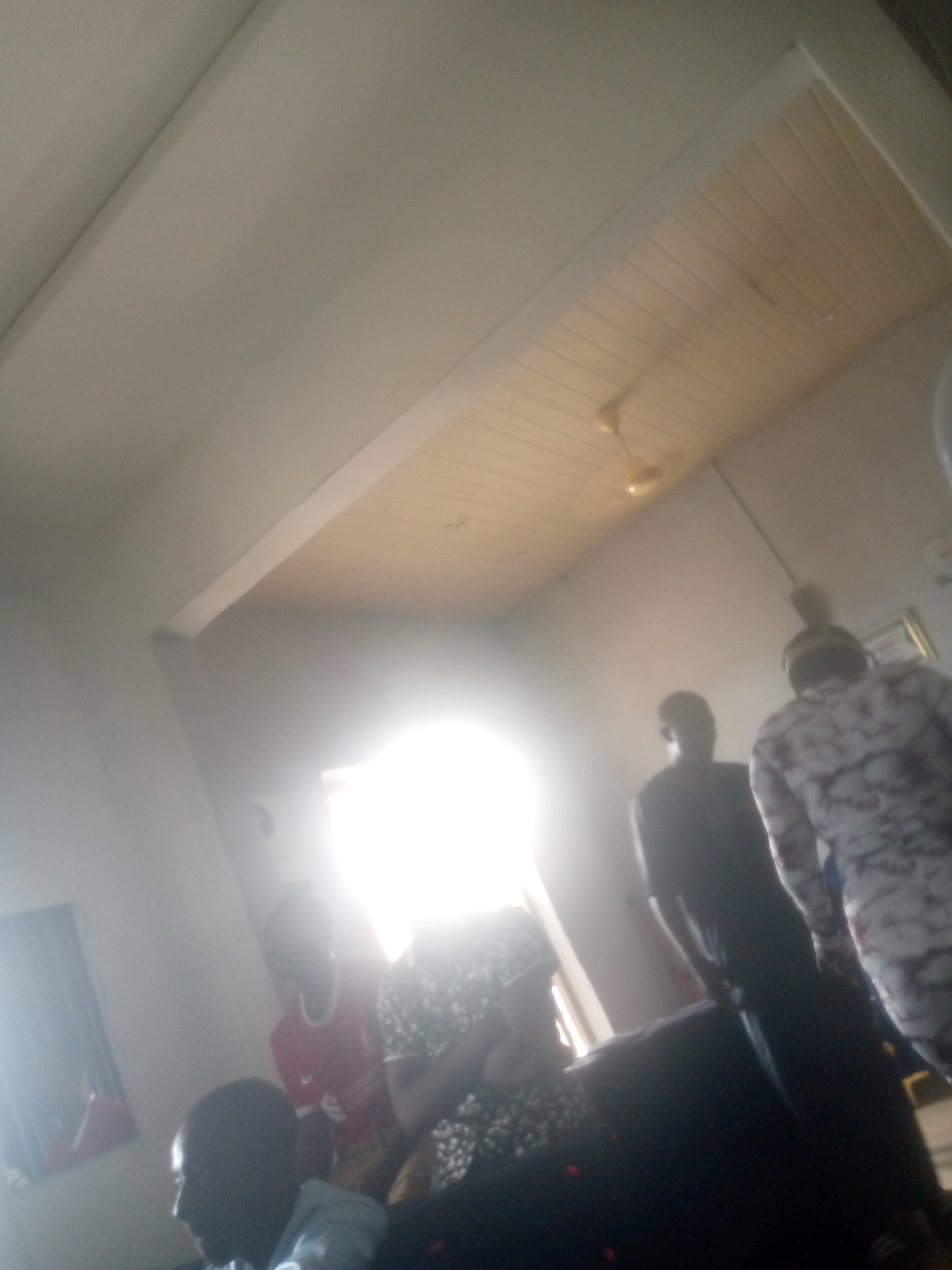 In an indoor residential setting, possibly a living room, in Jalingo, Nigeria, several individuals are gathered. A strong light source emanates from an opening, likely a window or doorway, obscuring details in the background and creating a bright, hazy atmosphere. The ceiling, constructed of light-colored wooden planks, is visible.
In the foreground, a person wearing a light-colored shirt is partially visible, facing left. To their right, another individual in a dark shirt is seen from the chest up, also facing left. Further back, a person in a dark patterned top is standing. To the left of this individual, a person in a red shirt is visible.
The overall image quality is poor due to low light and blurriness, making precise identification of features and actions challenging. The environment appears to be a domestic space, with furniture hinted at by dark masses in the lower right portion of the frame.