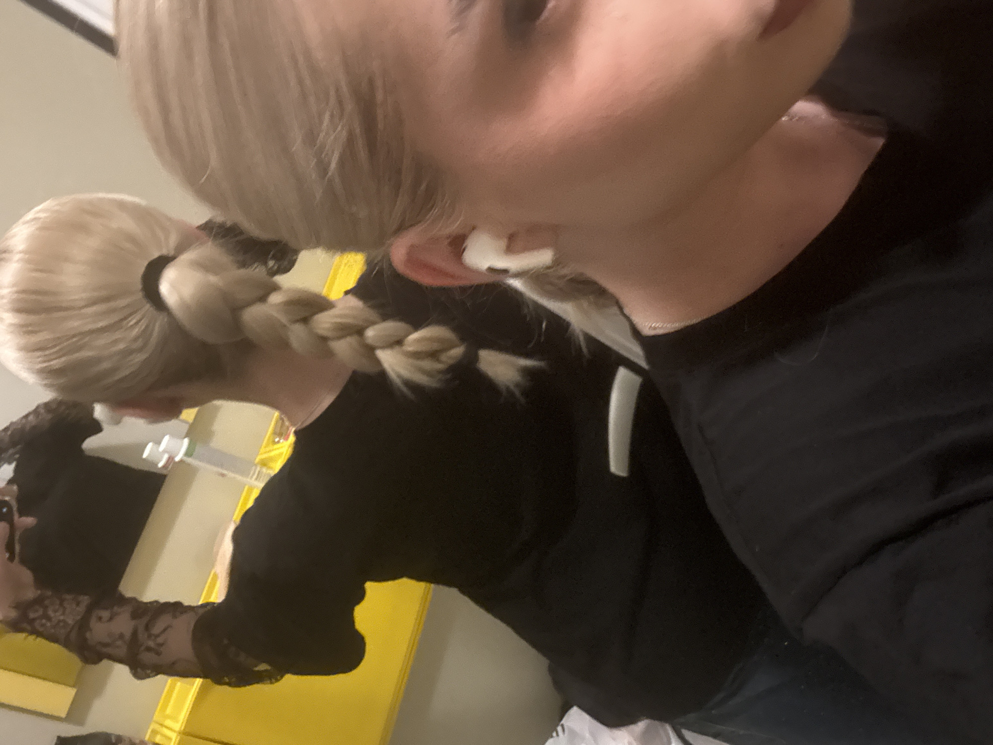 The image, taken in Sofia, Bulgaria, features a close-up, angled view of a person with blonde hair and fair skin. The subject is partially visible, seen from the side, with their head tilted. Their blonde hair is pulled back into a high ponytail, secured with a black hair tie, and then intricately braided down their back. A white earbud is visible in their right ear.
The person is wearing a black long-sleeved top. In the background, a reflection in a mirror shows the full upper body of the same individual. In the reflection, the person is seen holding what appears to be a smartphone in their left hand, indicating they are likely taking a mirror selfie. The reflected arm reveals a distinct black lace pattern on the sleeve, which adds a decorative element to the otherwise solid black top. A white vertical stripe is also faintly visible on the side of the reflected figure's black garment.
The setting appears to be indoors, with a bright yellow wall or surface visible in the background of the reflection. The lighting suggests artificial illumination. No specific time of day or emotional state can be discerned, and there is no visible text in the image.
