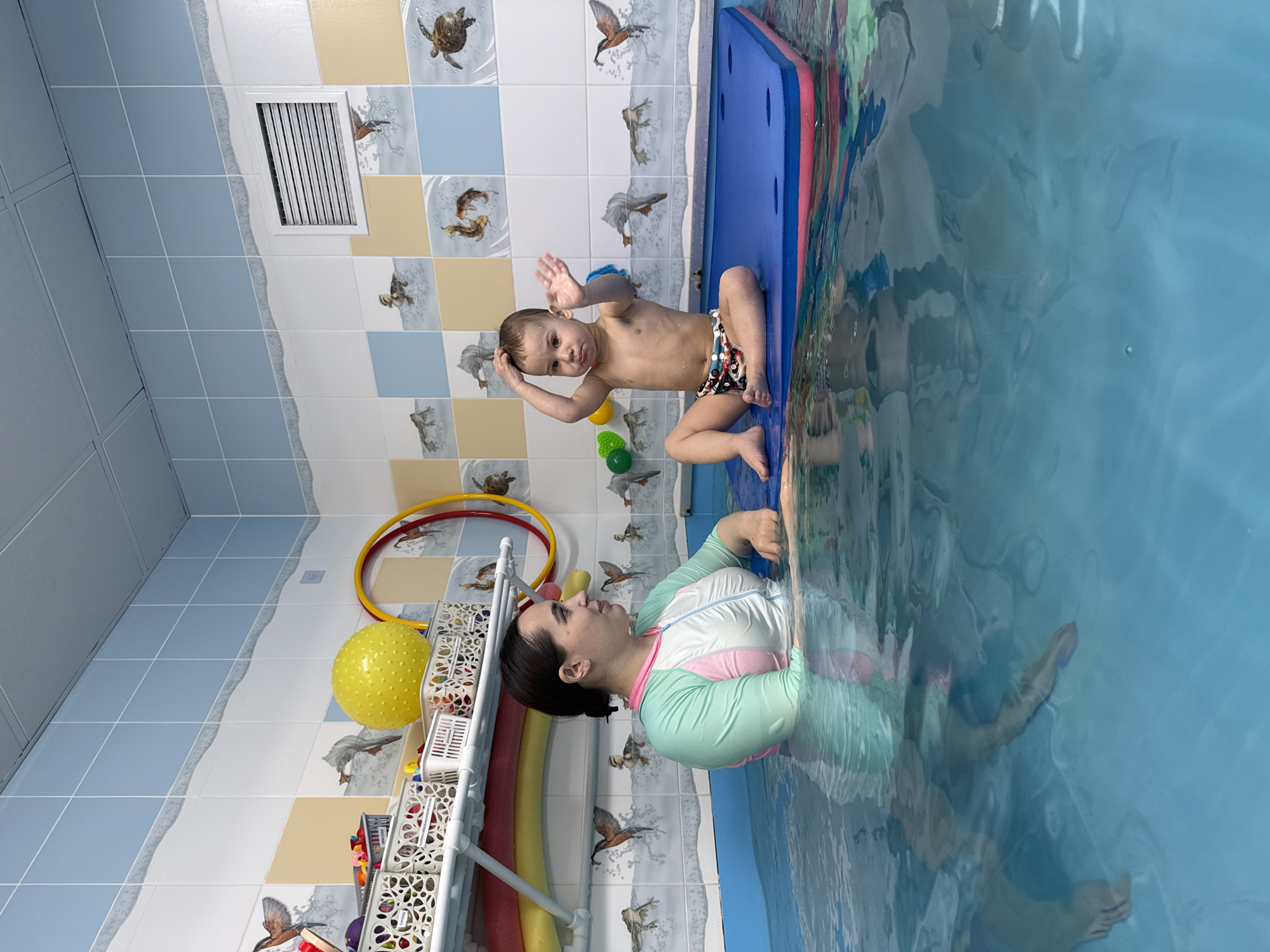 **Scene:** Indoor swimming pool in Rusanovo, Russia.
**People:** A young child and an adult female.
**Subjects:** The child is sitting on a blue and red floating pad, while the woman is in the water near the edge.
**Objects:**
* Swimming pool filled with blue water
* Blue and red floating pad
* Colorful patterned tiles on the walls with bird and turtle motifs
* White ventilation grate on the upper wall
* White shelving unit with colorful toys and balls, and a yellow hula hoop
* Yellow and green balls
**Actions:**
* The child has one hand on his head and one raised.
* The woman is in the water looking at the child.
**Interactions:** The woman is supervising the child who is sitting on the floating pad.
**Environment:** Brightly lit indoor swimming pool area with colorful decorations appropriate for children.