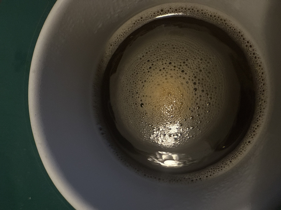 The scene, located in Arthès, France, presents a top-down view into a white or light grey ceramic mug. The mug contains a dark brown liquid, consistent with coffee, which is largely covered by a layer of fine, off-white froth. The froth displays a dense aggregation of small bubbles, arranged in a circular, somewhat concentric pattern across the liquid surface, with a slight visual density towards the center. A prominent, elongated reflection of an external light source, appearing bright white, is visible on the upper-left quadrant of the liquid, interacting with the froth and the underlying liquid. The upper inner rim of the mug is visible, and the mug rests on a dark green surface, portions of which are discernible at the top and top-left edges of the image. No individuals, specific actions, or interactions are depicted.