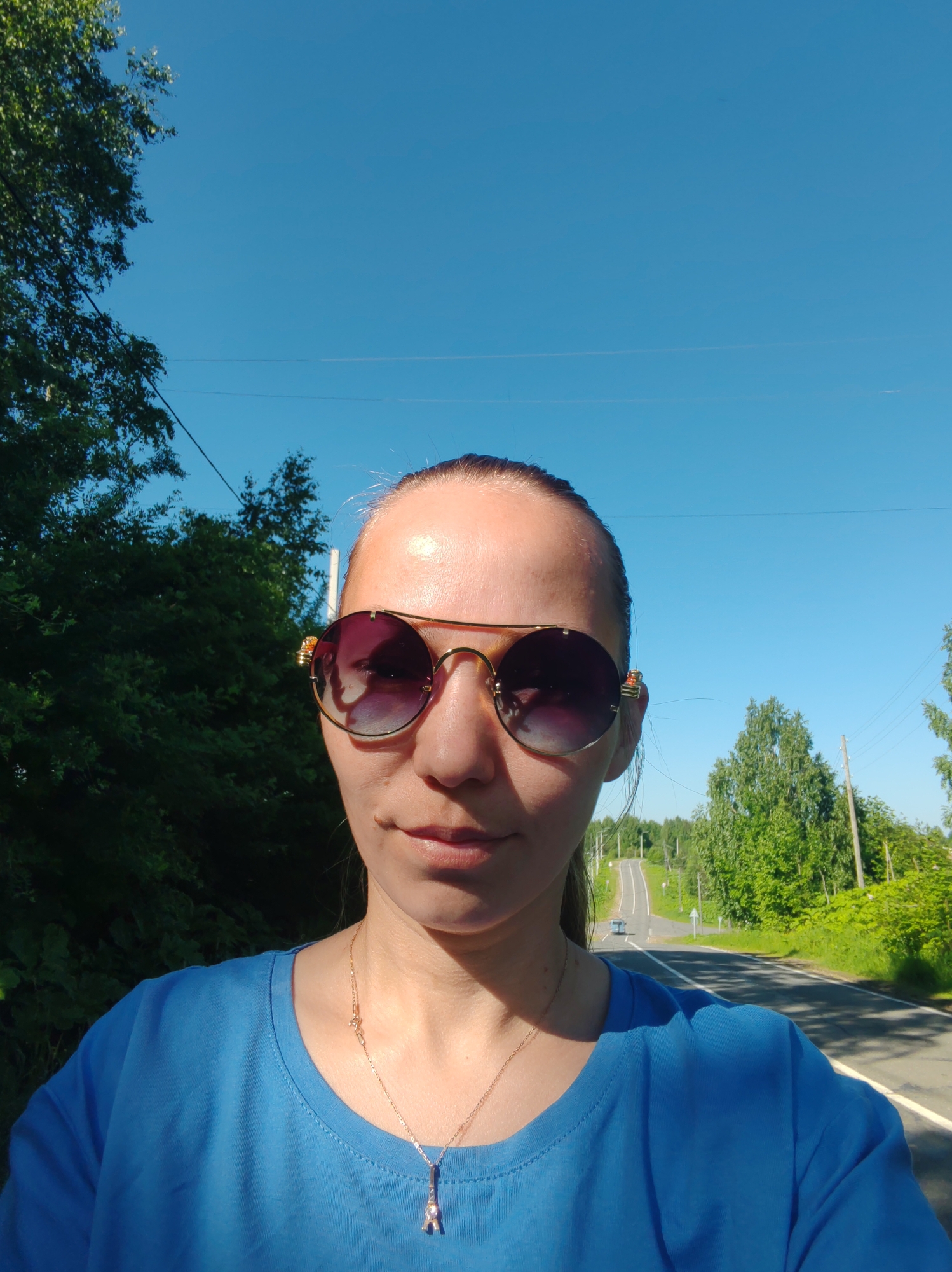 A woman in sunglasses and a blue t-shirt is taking a selfie outdoors. She has her hair pulled back. The woman is standing on a road with trees and power lines on either side. The road appears to be in a rural or suburban area. The sky is clear and blue. The location is specified as Chukhlem, Russia.