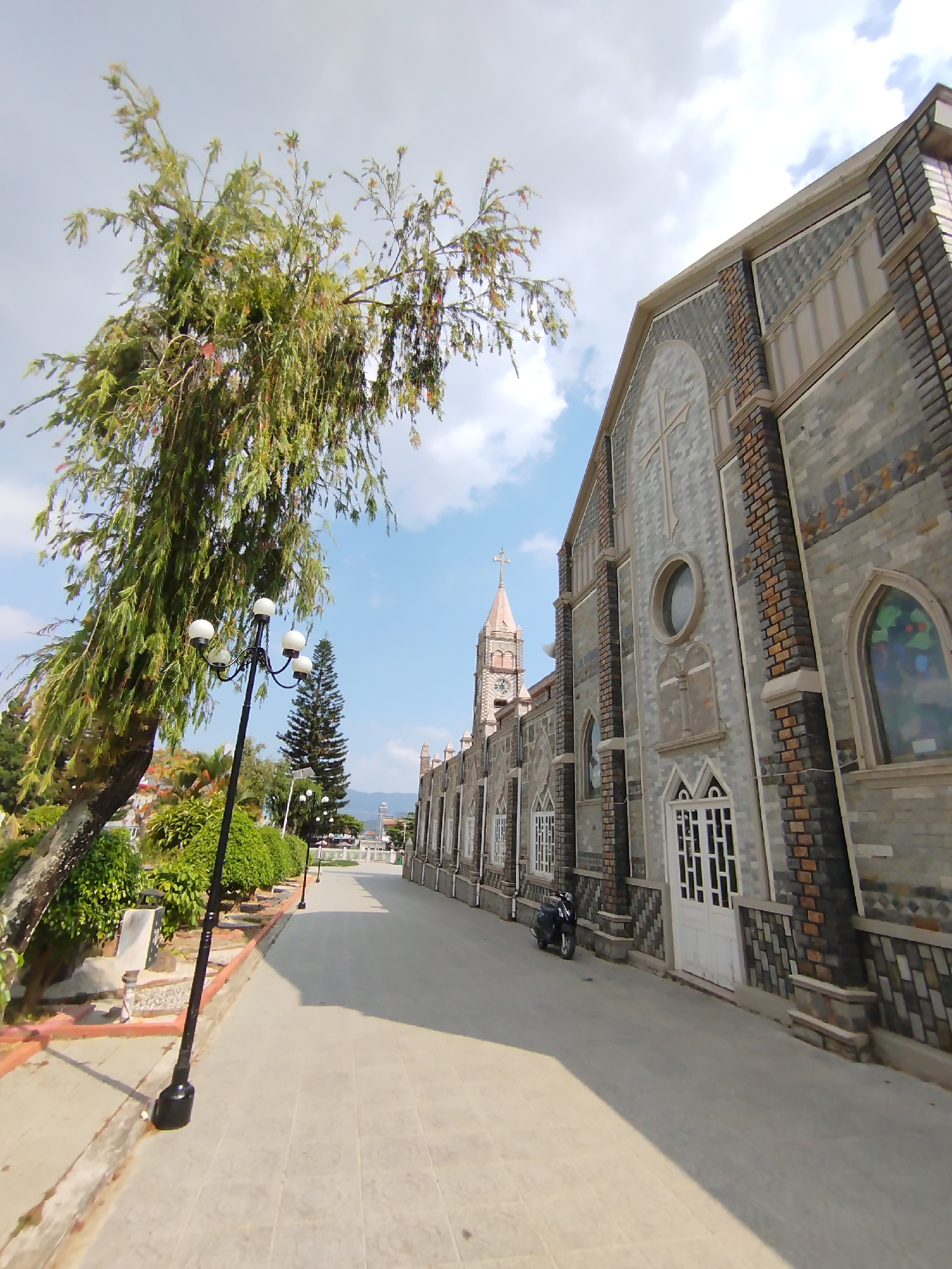 The image captures a serene outdoor scene featuring a prominent church in Lạc Viên A, Vietnam, under a partly cloudy blue sky. The main subject is a large, stately building with architecture reminiscent of a church, characterized by patterned brickwork and grey stone facades. Its side features a large cross emblem, circular windows, and tall arched windows, one of which appears to have stained glass. Further down the street, a clock tower with a pointed spire topped by a cross is visible, indicating the main entrance or bell tower of the church.
In the foreground on the left, a large tree with long, drooping green leaves frames the scene. A black street lamp with four spherical white lights stands beneath the tree. A paved pathway runs alongside the tree and the church, leading into the distance where more street lamps and greenery line the route. On the path next to the church, a single dark-colored motorcycle is parked. The overall setting appears well-maintained, with neatly trimmed bushes and additional trees dotting the landscape.
The time of day is bright daylight, suggested by the clear illumination and the presence of shadows cast by the tree and street lamp. No people are visible, contributing to a tranquil and quiet atmosphere. There is no discernible text within the image.